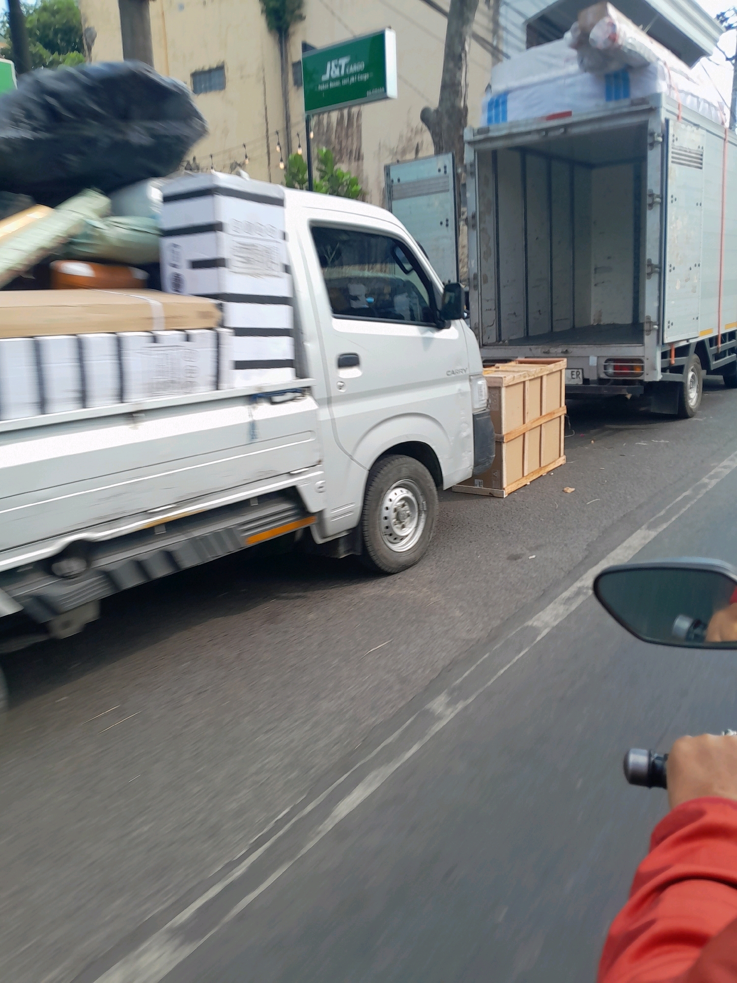 In the city of Ardirejo, Indonesia, on a paved road with white lane markings, two trucks are shown. The first truck, a white cargo truck, is heavily laden with boxes and other items, including a large black bag. It appears to be moving or has recently stopped. To its right, a larger, silver box truck is parked with its rear doors open, revealing an empty interior. In front of the white truck and to the right, a wooden crate is positioned on the road. The scene appears to be an outdoor, daytime setting. A hand in a red jacket is visible on the right side of the frame, appearing to be on the handlebar of a motorcycle, suggesting a person on a motorcycle is observing the scene. The background includes buildings and trees.