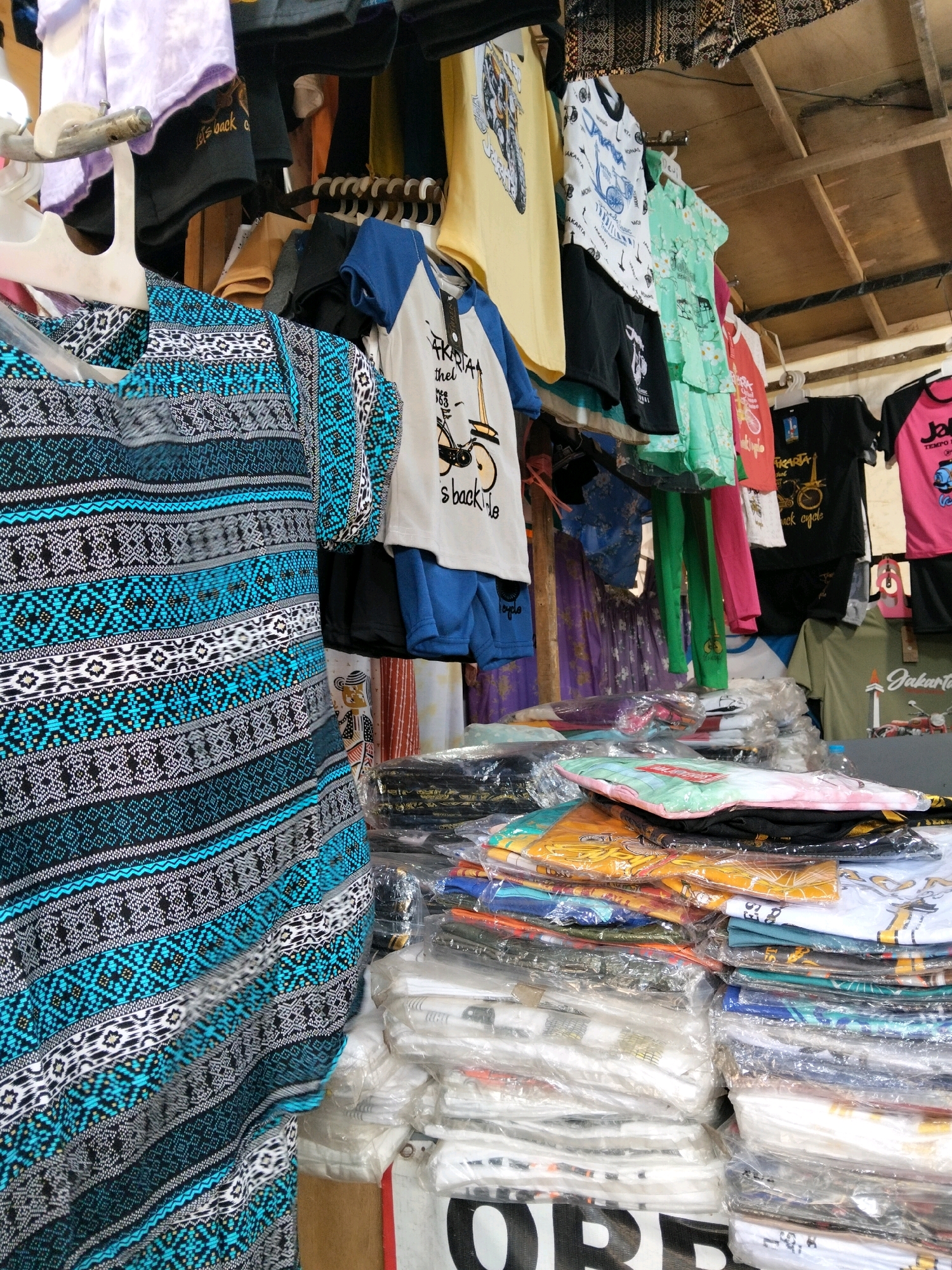 This image depicts a close-up view of a market stall selling clothing in Central Jakarta, Indonesia. The stall displays a variety of t-shirts and clothing items hanging and stacked. In the foreground, a dress with a blue, black, and white patterned fabric is prominently displayed. Behind it, a selection of t-shirts with graphics and text, including one with the word "Jakarta" and imagery of a bicycle, are visible. The stall appears to be covered by a wooden structure, suggesting an outdoor or semi-outdoor setting. The overall scene conveys a typical street market environment.