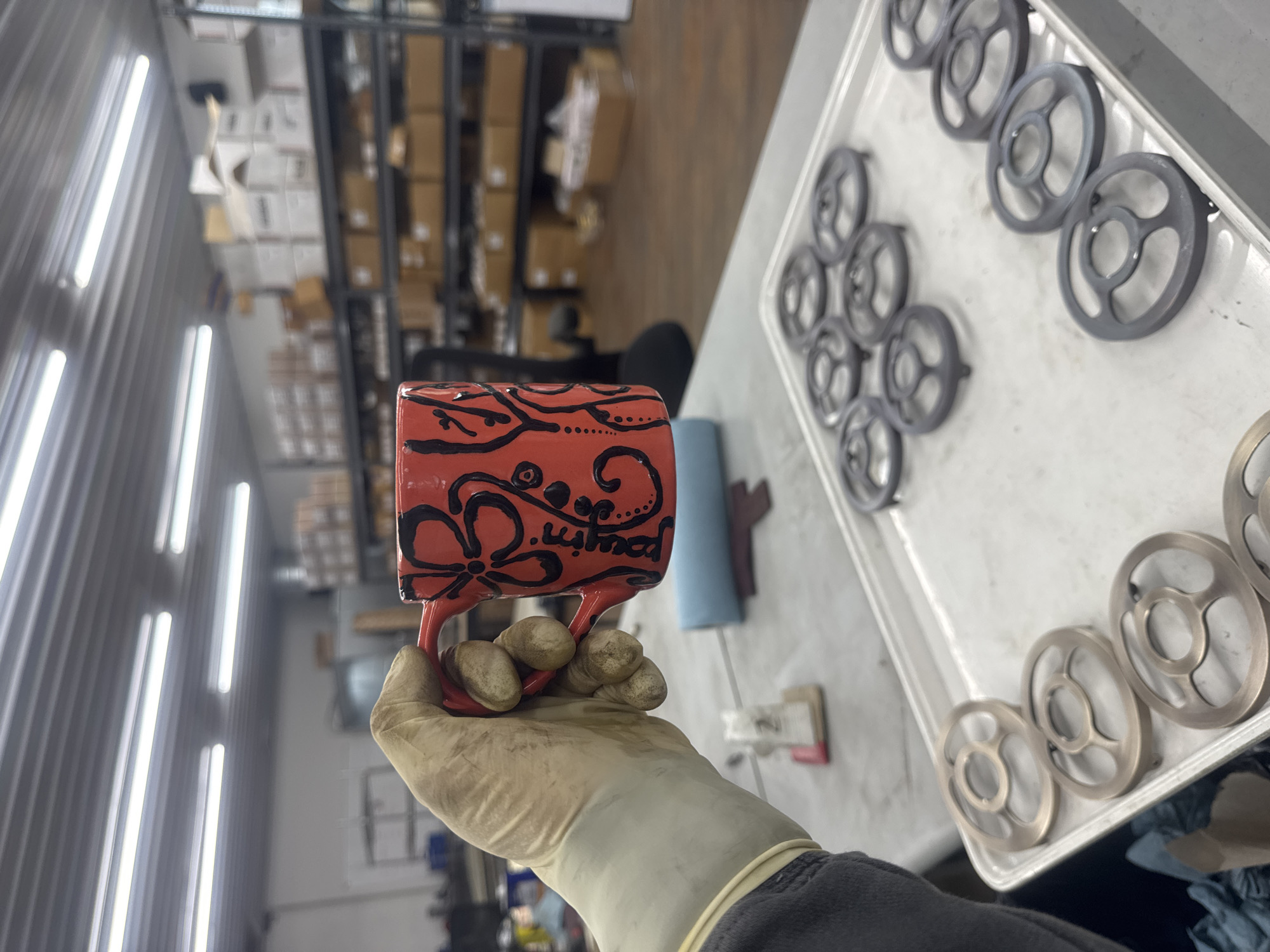 The image shows a hand wearing a beige glove holding a red mug decorated with black floral designs. The mug has a handle and is decorated with swirling lines and flower-like shapes. Some text, possibly a name, appears on the mug in black ink, though it is difficult to read clearly. In the background, a white tray filled with circular metal objects is visible, arranged in neat rows. Beyond the tray, shelves filled with boxes are seen in a warehouse or storage setting. The lighting suggests an indoor environment, possibly a workshop or industrial space, with bright fluorescent lights visible on the left side of the frame. The overall scene appears to be a still life composition, with a focus on the mug and the surrounding objects.