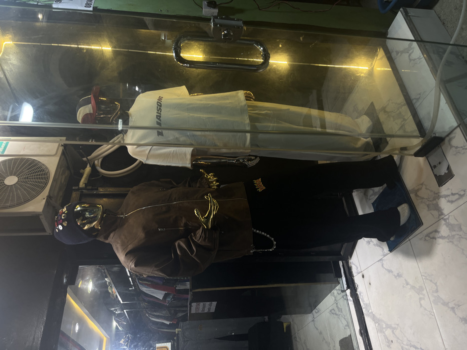 This image depicts a retail storefront located in Iranla, Nigeria, displaying two mannequins through a glass partition. The mannequin on the left has a gold-toned head and hands, wearing a dark blue beanie embellished with multi-colored circular studs, a brown zippered jacket, and black pants with a gold crown logo near the left hip, from which a silver chain dangles. This mannequin stands on a blue mat, wearing white footwear. The mannequin on the right, also with a gold-toned head and hands, wears a red and white baseball cap, an off-white short-sleeved t-shirt bearing the word "LIAISON" in dark grey letters, and matching off-white pants. This mannequin stands on a dark mat, wearing dark footwear.
Behind the mannequins, visible through another glass partition, is the store interior with racks of clothing and bright overhead lighting. Above the mannequins, a white Hisense brand air conditioning unit is mounted, with exposed electrical conduits and wiring. The exterior floor is covered with white marble-patterned tiles. To the right of the glass door, a green wall is visible, featuring a vertical yellow LED strip light, a chrome door handle, and a small white sign with partial text "0814..." affixed to it.