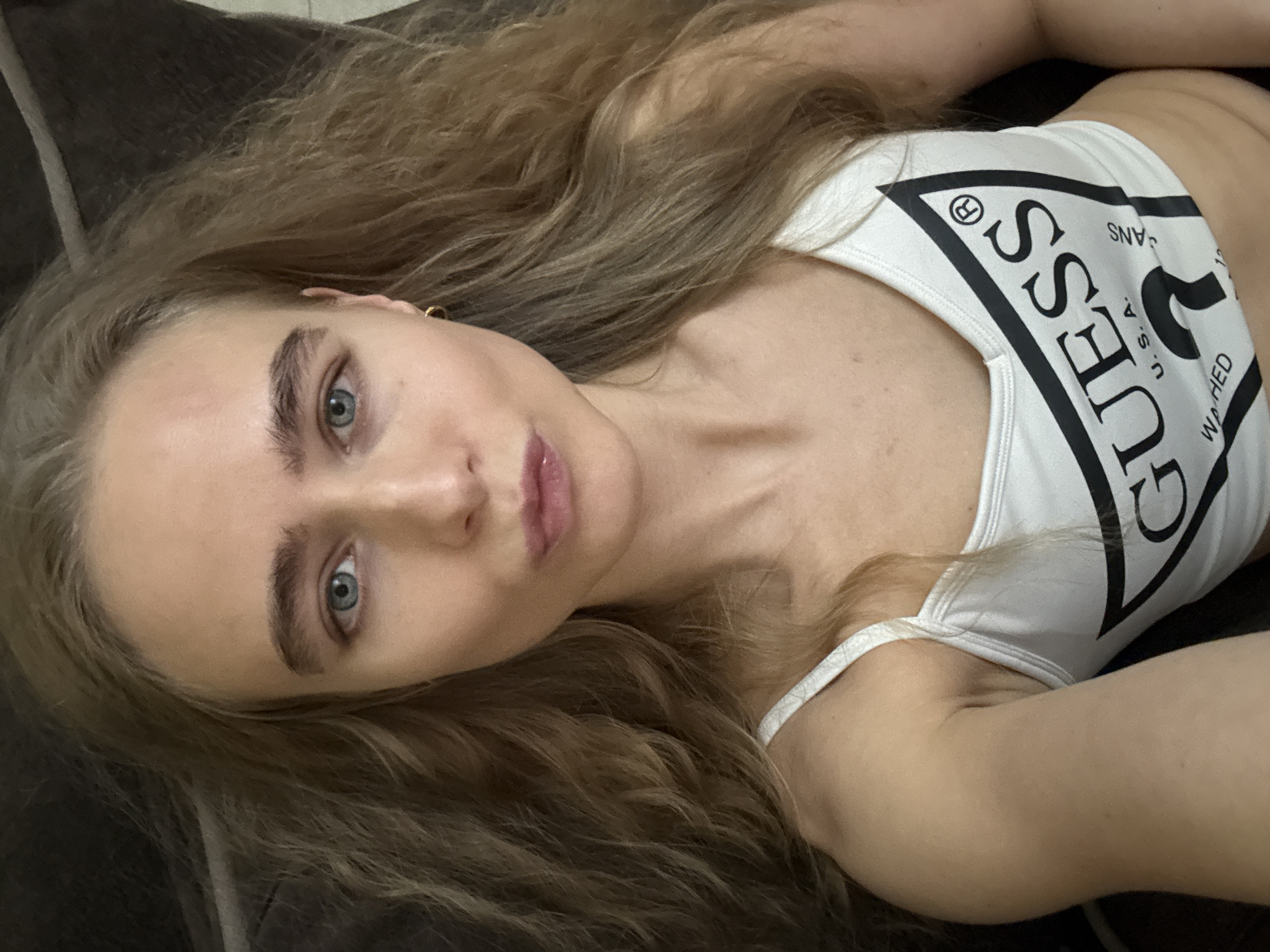 A single female individual is depicted in a close-up, top-down selfie. The location is an indoor setting, likely a private residence, in the city of Unknown, Unknown. The subject has long, wavy, light brown hair, fair skin, and blue eyes. Her lips are slightly pursed. She wears a small gold hoop earring in her visible right ear. She is dressed in a white sleeveless crop top with thin straps, featuring a prominent black triangular "GUESS U.S.A." logo on the chest. The background is dark and out of focus, providing no additional specific environmental details.