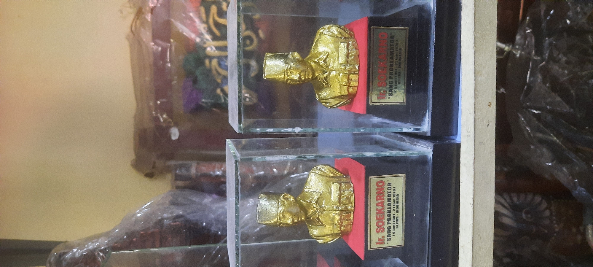 Two identical busts of Ir. Soekarno, a key figure in Indonesian history, are displayed in separate glass enclosures. Each bust is golden in color and sits atop a red pedestal, which is mounted on a black base. A gold plaque is affixed to the front of each black base. The left plaque reads "Ir. SOEKARNO (6 Juni 1901 - 21 Juni 1970) BLITAR - INDONESIA". The right plaque is partially obscured but appears to have identical text. The busts are displayed on a shelf in what appears to be a shop or collection. Behind the busts, out-of-focus items are visible, including what looks like packaging and other decorative objects. The lighting suggests an indoor environment, possibly with natural light supplemented by artificial sources. The presence of the Blitar, Indonesia inscription on the plaque strongly suggests this location is within or related to the city of Blitar.