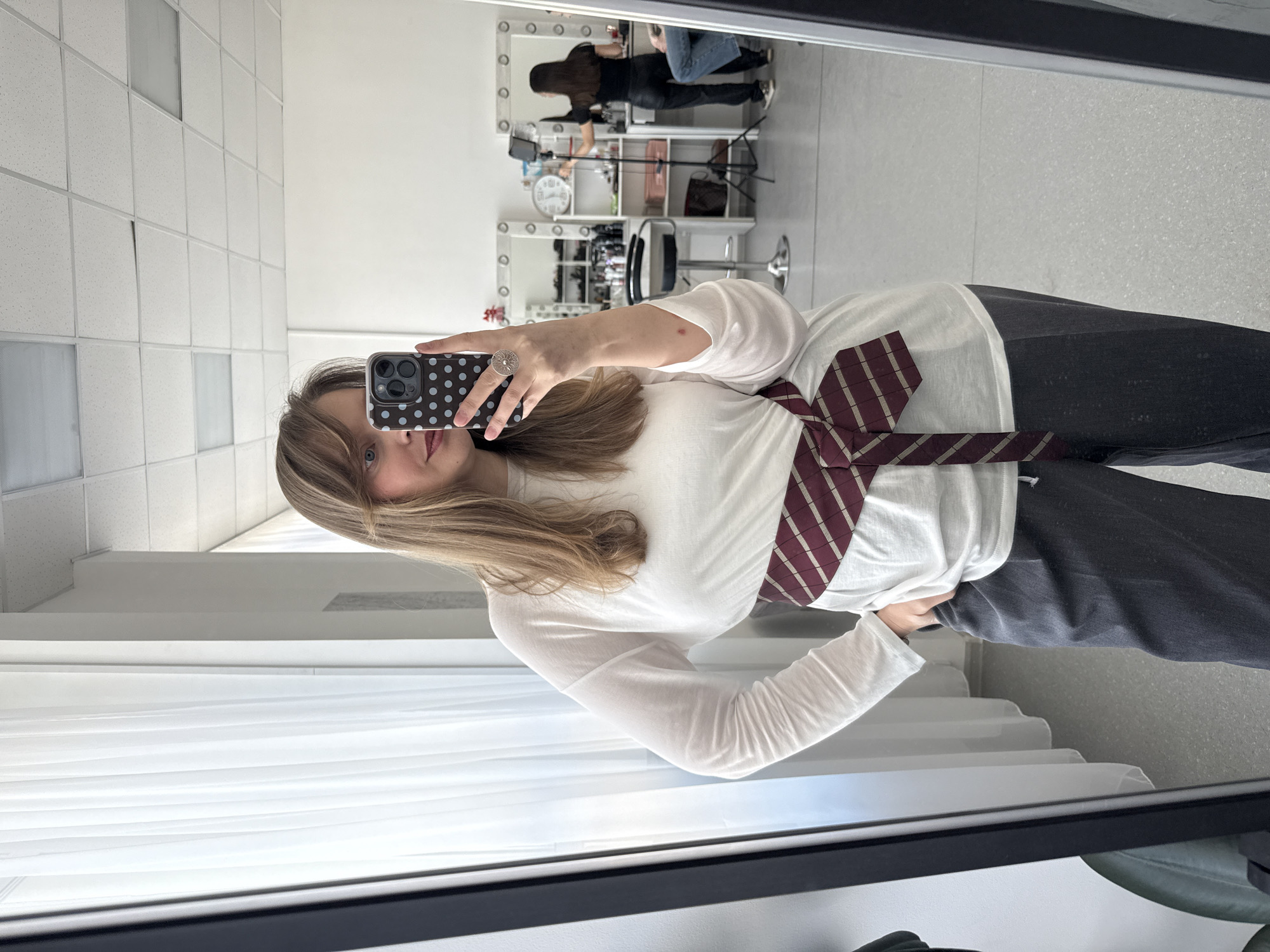A woman is taking a selfie in a mirror. She is wearing a white long-sleeved shirt and grey pants. A burgundy and white striped tie is around her waist. The reflection in the mirror shows a room with a vanity table, a clock, and other people. The location is likely indoors, possibly a dressing room or salon. The city is not discernible from the image.