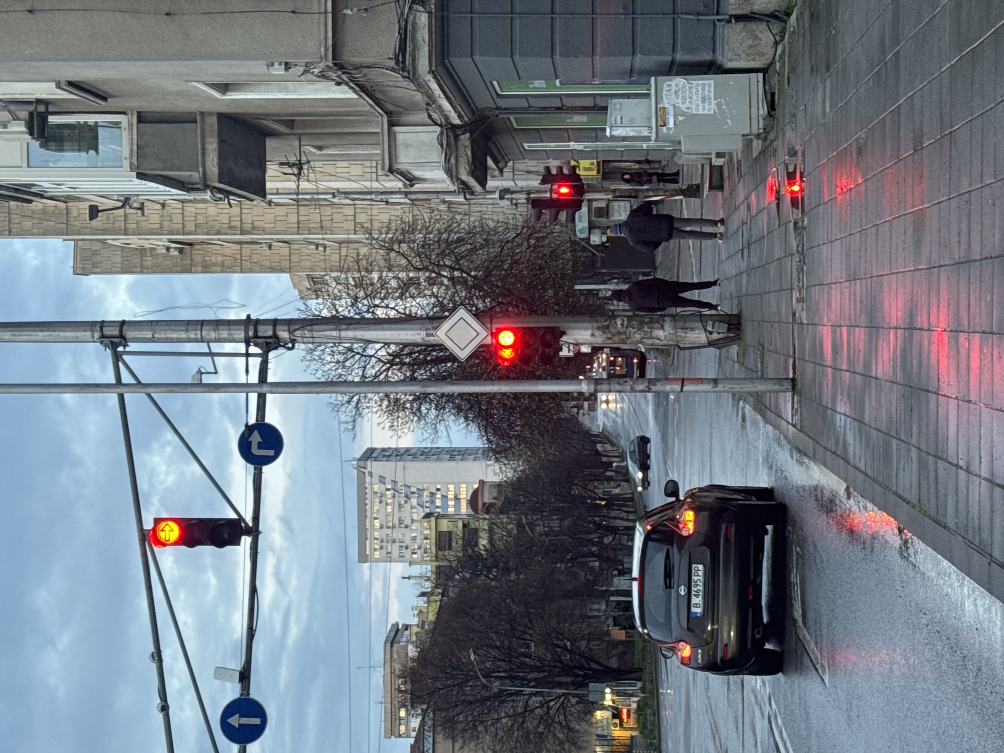A street scene in Burgas, Bulgaria, shows a wet road surface reflecting red traffic lights. A dark-colored Nissan vehicle with license plate "B 4695 PP" is stopped in the right lane. Above the car is a row of trees and buildings on the street. People are walking on the sidewalk. Three sets of traffic lights are visible; all displaying red. A main road sign indicating "Priority road" is also visible above the middle traffic light. The sky is overcast. Several buildings are situated in the background with lit windows.