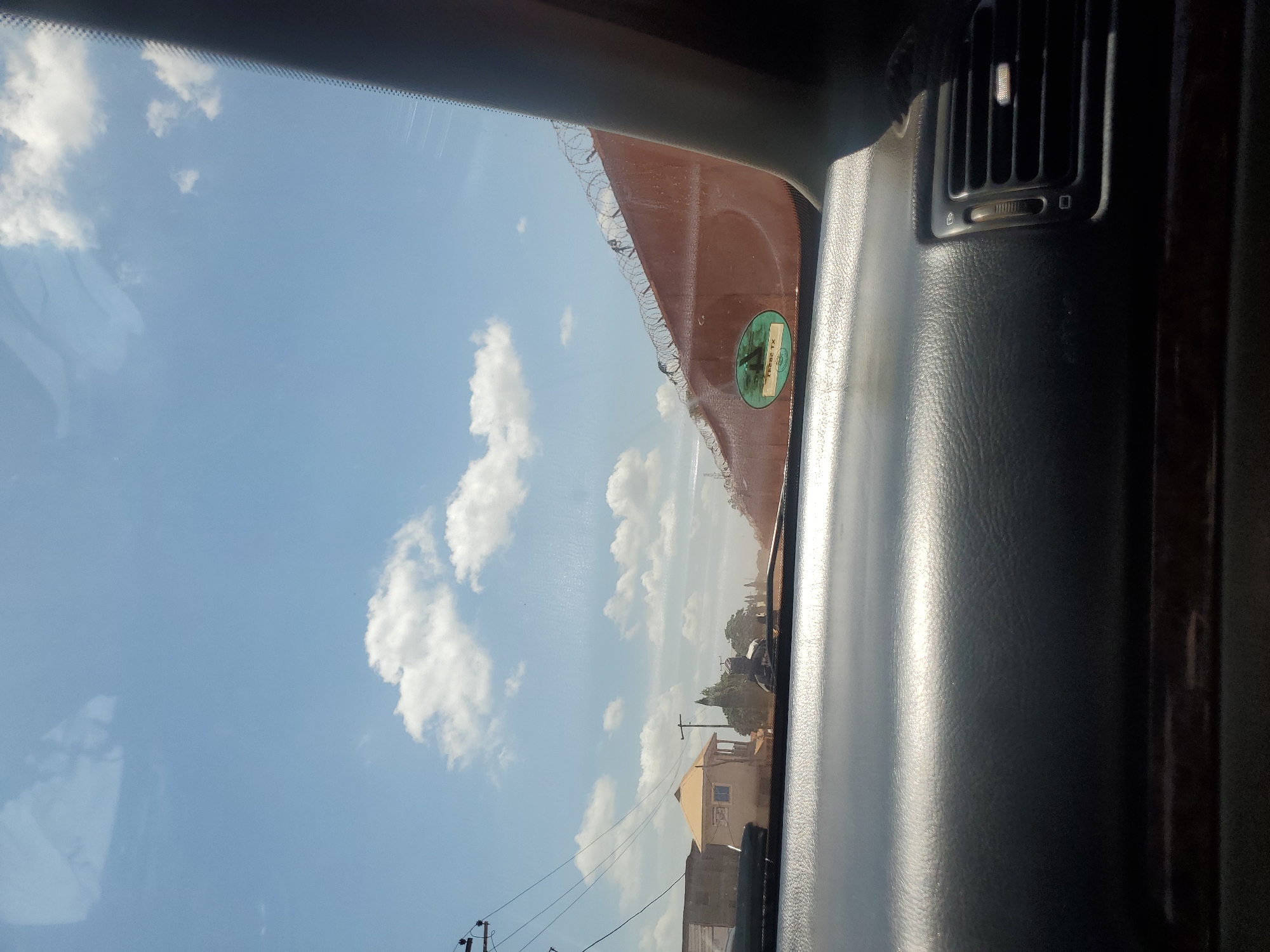 The image depicts a bright daytime scene in Yelwa, Nigeria, viewed from inside a vehicle through its windshield. The foreground features a dark-colored dashboard, reflecting sunlight, with a visible air vent on the far right. The sky is clear blue with scattered white cumulus clouds.
On the left side of the exterior view, utility poles with multiple overhead wires are present. A light-colored building with an orange-brown pitched roof and a veranda stands, with a plainer concrete structure visible behind it, along with distant trees. A dark, indistinct shape, possibly a person on a motorcycle or a stationary object, is visible in the middle ground, partially obscured by reflections on the windshield.
On the right side of the exterior view, a long, reddish-brown wall topped with razor wire extends into the distance, indicating a secure perimeter. A green oval sticker with the text "TRD82 TX" is affixed to the interior of the windshield, partially overlapping the view of the wall. The road surface in the middle distance appears unpaved and dusty. Reflections, possibly of the vehicle's interior or the sky, are visible on the upper left portion of the windshield.