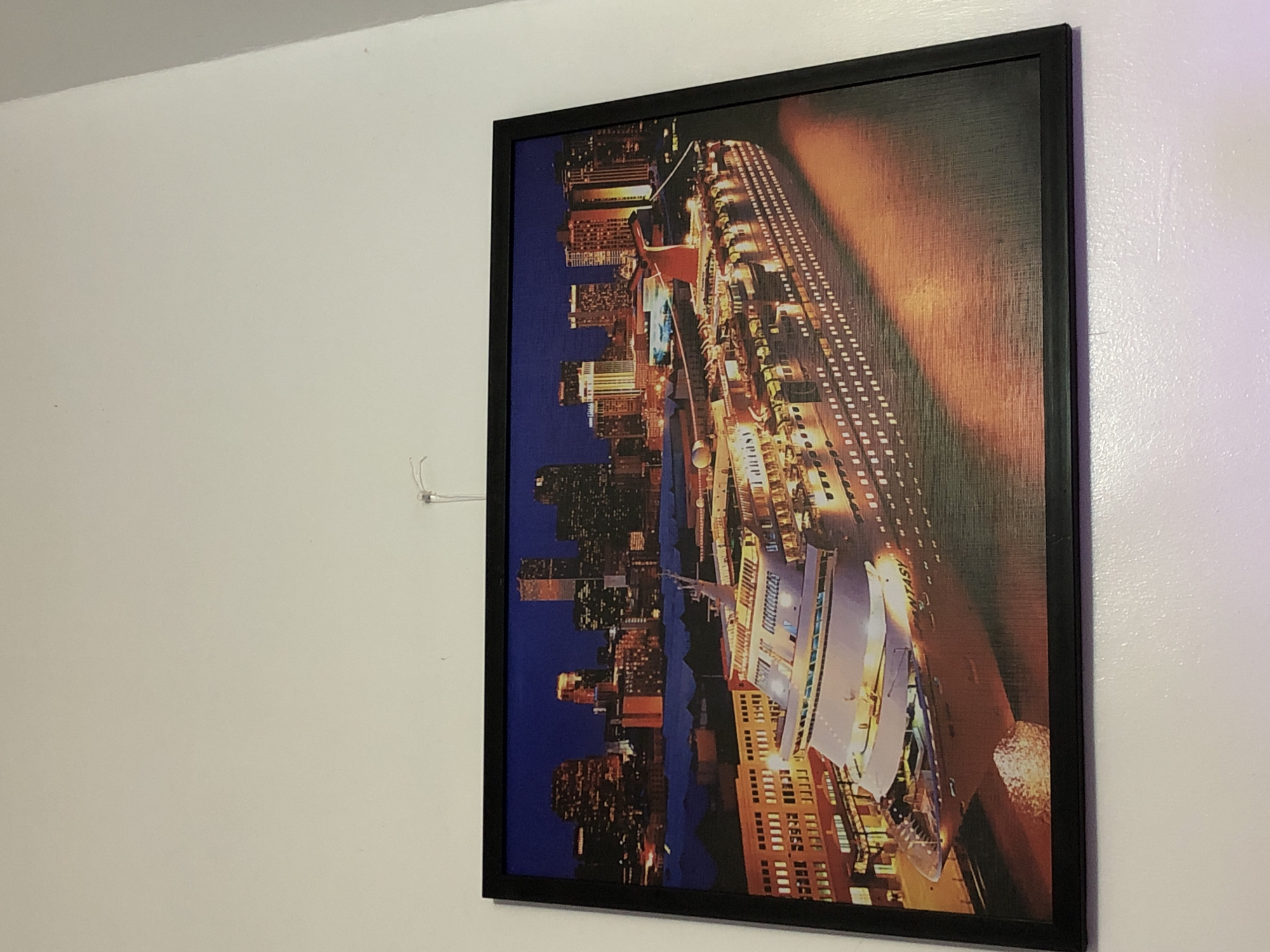 An image displays a framed picture hanging on a white wall, rotated ninety degrees counter-clockwise. The picture features a vibrant night scene of a large, illuminated cruise ship or ferry in the foreground, with a bustling cityscape stretching behind it. Numerous tall buildings with glowing windows are visible across the dark blue night sky, suggesting a major metropolitan area with a significant port.
The cruise ship is brightly lit, showcasing multiple decks and structures. Part of the name "FANTASY" is visible in white letters along the side of the ship. A striking warm, orange-toned diagonal light effect spans the upper right portion of the artwork, adding to the dramatic nighttime ambiance. The water in the harbor appears dark, reflecting some of the city lights.
The frame of the picture is black and has a thin profile. The wall to the left of the frame is plain white, with a subtle texture and some reflections. A small, white, translucent plastic hook or clip, with a thin wire or string attached, is visible on the wall to the left of the picture, positioned slightly above its center. The overall impression is a dynamic, well-lit urban waterfront at night, captured in an artwork.
