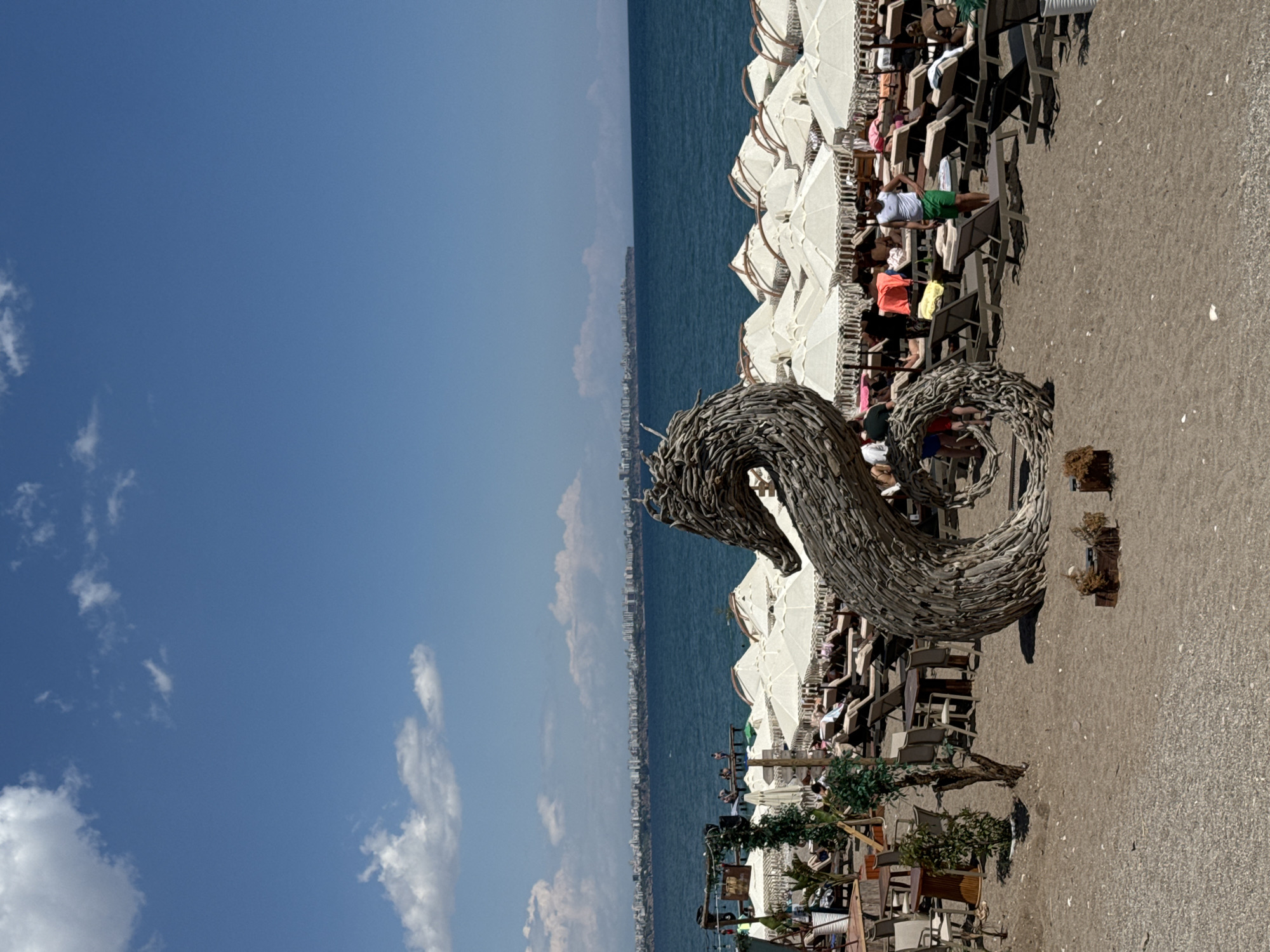 The image shows a beach scene in Konyaaltı, Turkey on a clear, sunny day. A large, driftwood sculpture of a dragon or sea serpent is positioned prominently on the sand. Behind the sculpture are rows of white sun umbrellas and beach chairs occupied by people, some appearing to be sunbathing or relaxing. In the background, the blue sea stretches towards a distant coastline dotted with buildings, suggesting a cityscape along the coast. The sky is bright blue with scattered clouds.