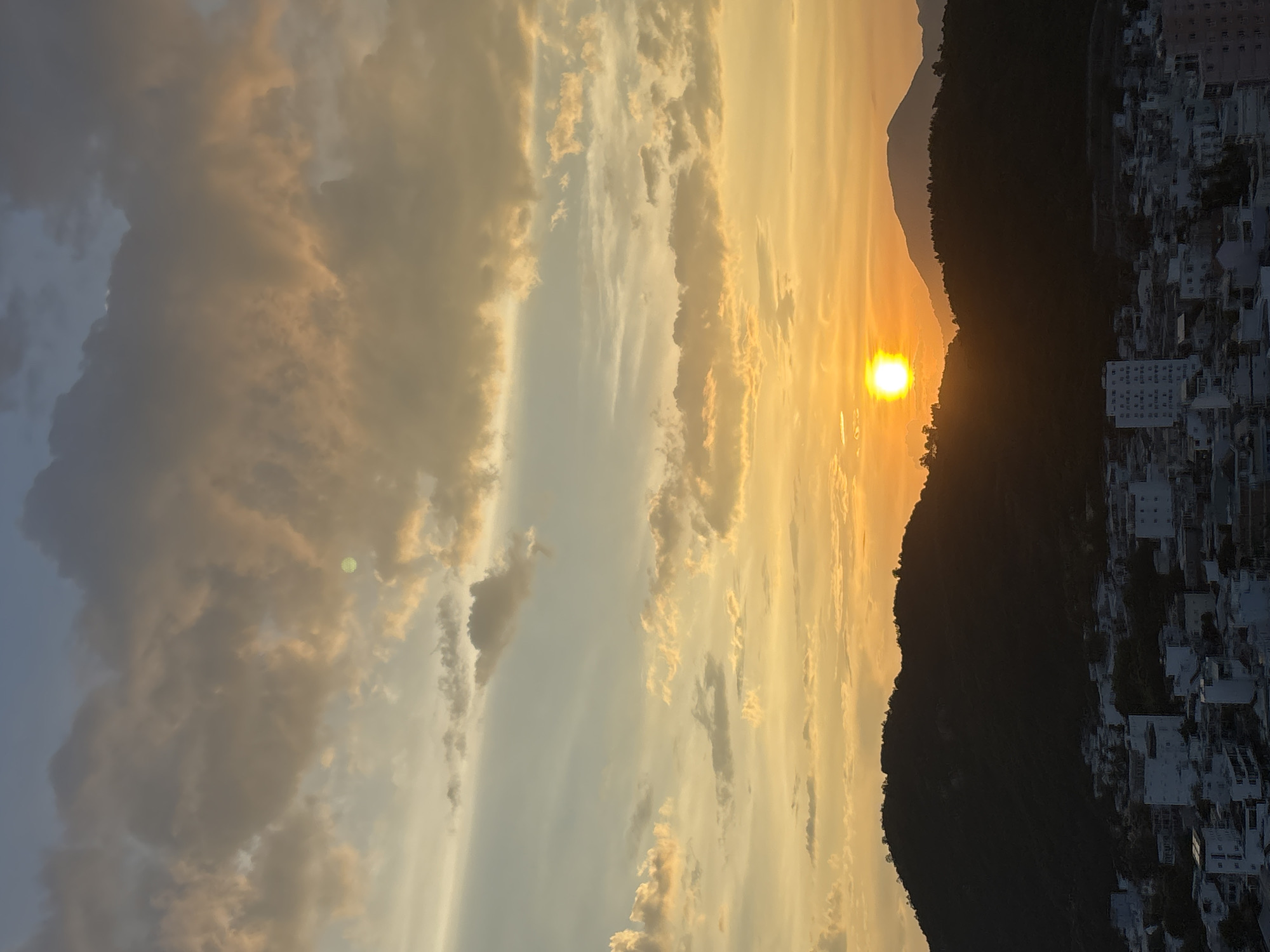 A wide shot captures a city at sunset. The sky is a gradient of soft blues and oranges, filled with scattered clouds tinged with golden light. The sun, a bright orange orb, is partially obscured by a dark, silhouetted mountain range that dominates the middle ground. The mountains are covered in dense foliage, and their peaks are dotted with small trees. In the foreground, a densely populated urban area is visible, with numerous buildings of varying heights and styles. The buildings are mostly white and light-colored, with some appearing to be residential or commercial structures. The overall scene is one of tranquility and natural beauty juxtaposed with urban development.