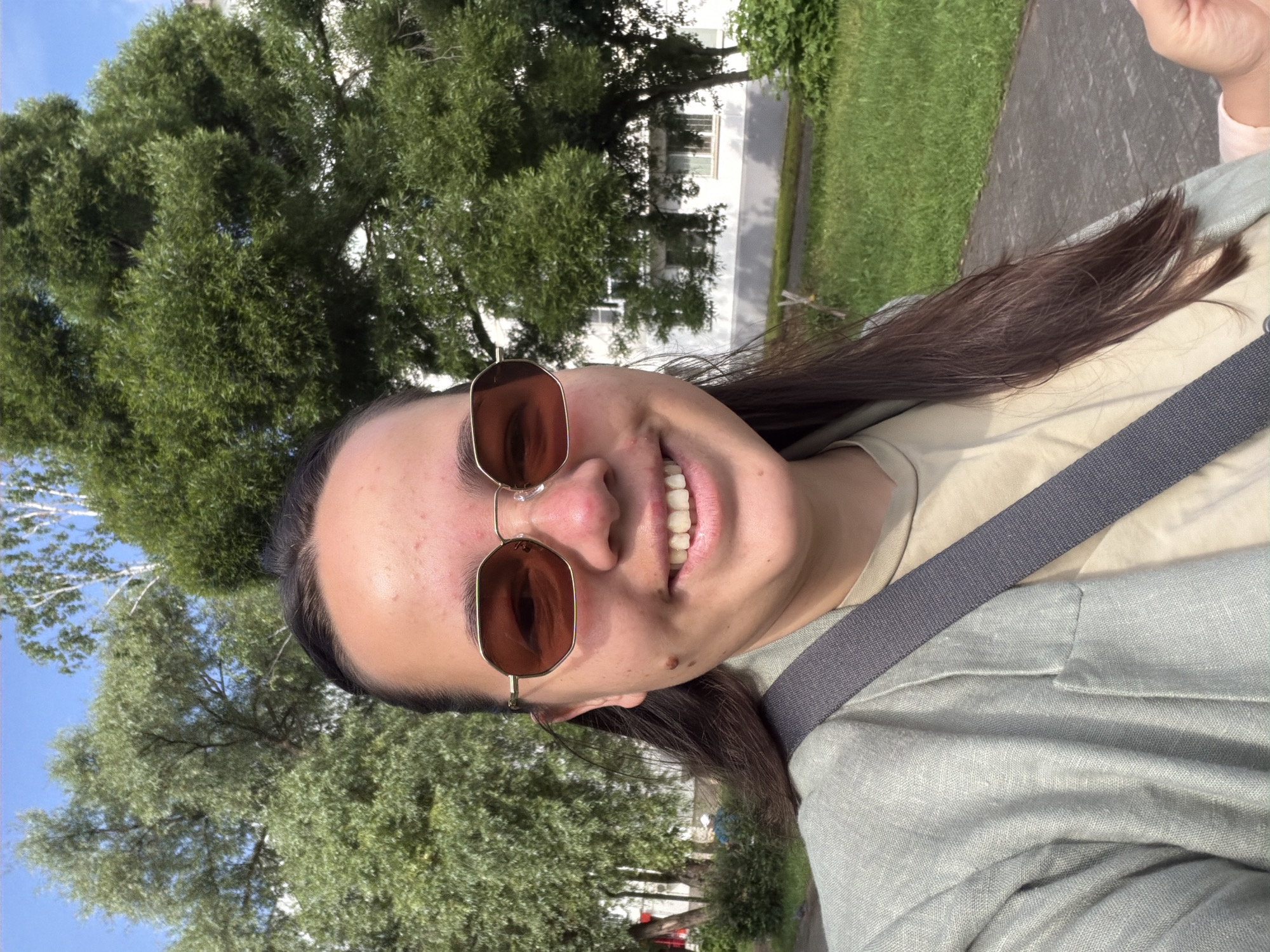 A young woman with dark hair, wearing sunglasses, a light green blazer, and a beige t-shirt, smiles at the camera. She has a dark mole on her left cheek. A gray strap crosses her chest. She is standing outdoors with lush green trees and a white building in the background, suggesting a residential area. The ground appears to be a paved walkway with grass to the side. The scene is likely in Chaikovsky, Russia, given the context. The lighting indicates it is daytime.