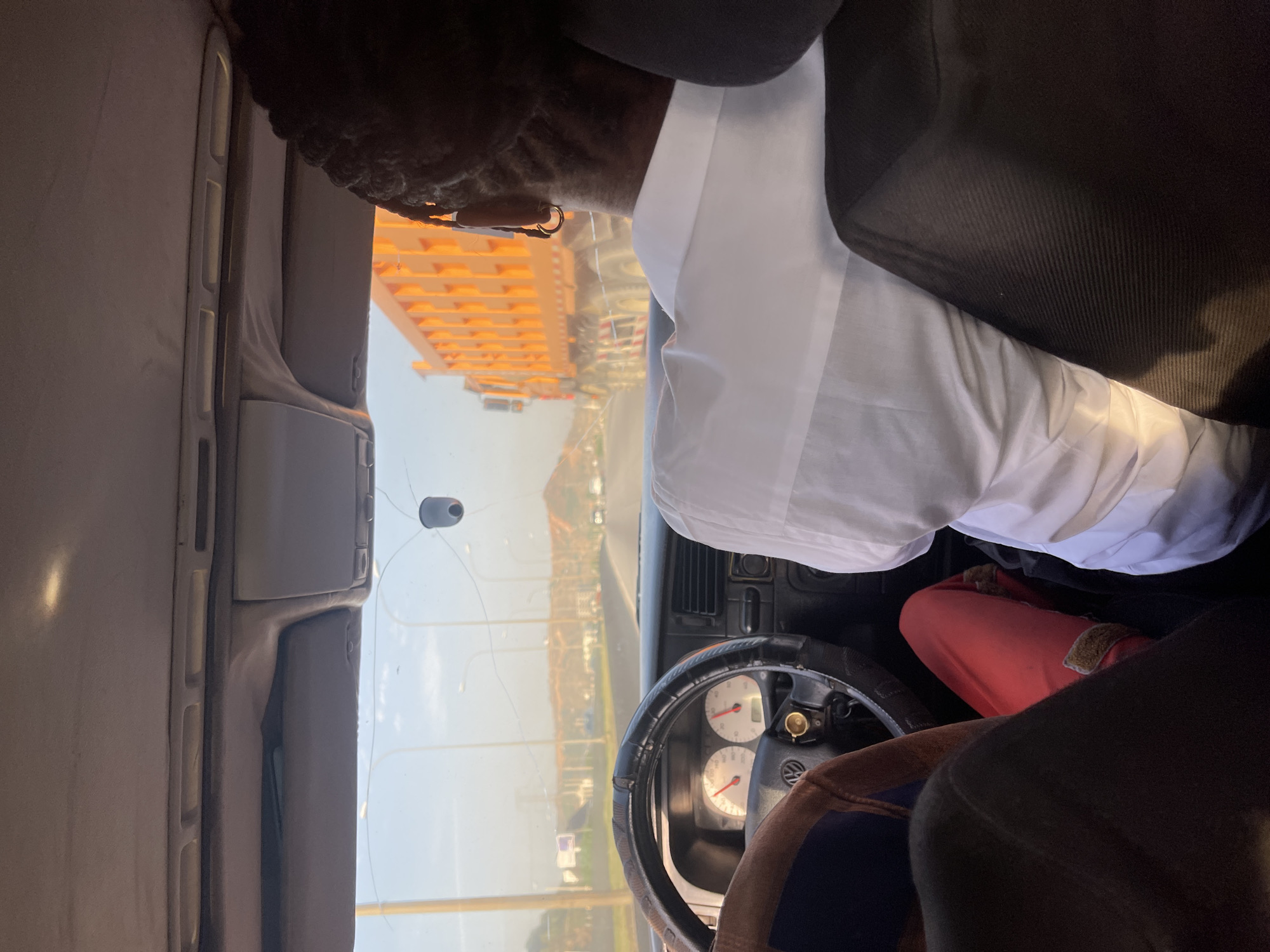 The image shows the interior of a vehicle, specifically the driver's seat. The viewpoint is from the passenger seat. A dark-skinned person with braided hair, wearing a white shirt and a black jacket, is partially visible in the driver's seat. The driver's hands are on the steering wheel. The dashboard is visible, with gauges and controls. The windshield has a crack. Through the windshield, a view of Jiru, Nigeria, is visible. An orange building is prominent in the background. The sky is light.