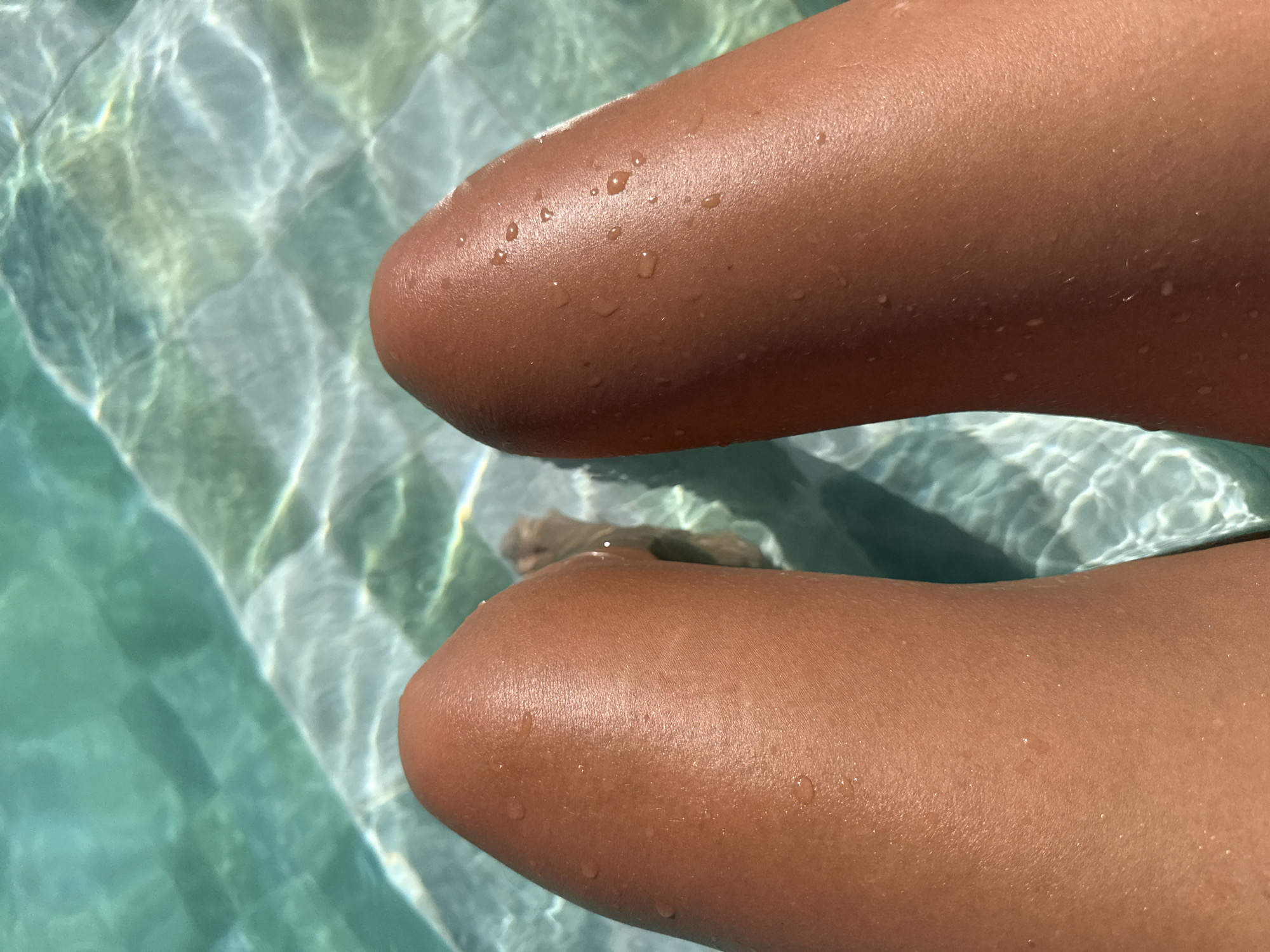 The image displays a close-up, top-down view of two human legs, exhibiting a tanned complexion, partially submerged in a swimming pool in Tivat, Montenegro. Numerous clear water droplets are visible on the sun-kissed epidermal surface of both legs. The pool water is distinctly turquoise, with patterns of refracted sunlight dancing across its surface and illuminating the submerged white or light-colored square tiles on the pool floor. A blurred outline of human feet is discernible further down in the water.