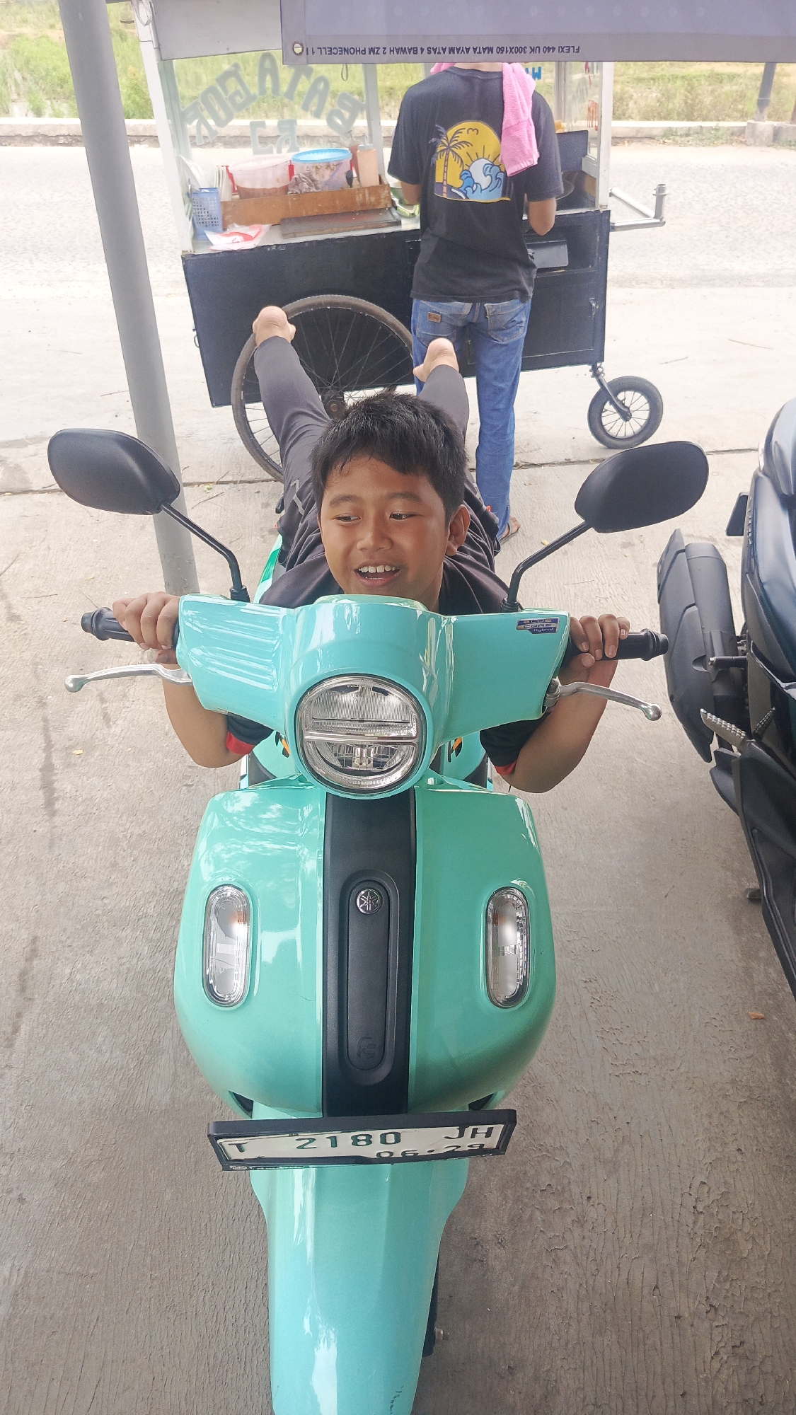 A young boy lies on a mint green scooter, holding the handlebars and smiling. His legs are extended behind him, with one foot resting on the wheel of a nearby food cart. The food cart, which appears to be mobile, has a clear plastic front displaying food items. Behind the cart, a person wearing a black t-shirt and blue jeans stands, with a pink towel draped over their shoulder. The scooter has a license plate with the number "T 2180 JH" and the date "06.29". The scene takes place outdoors, on a paved surface, with a hint of greenery in the background, suggesting a location in Parung Panjang, Indonesia.