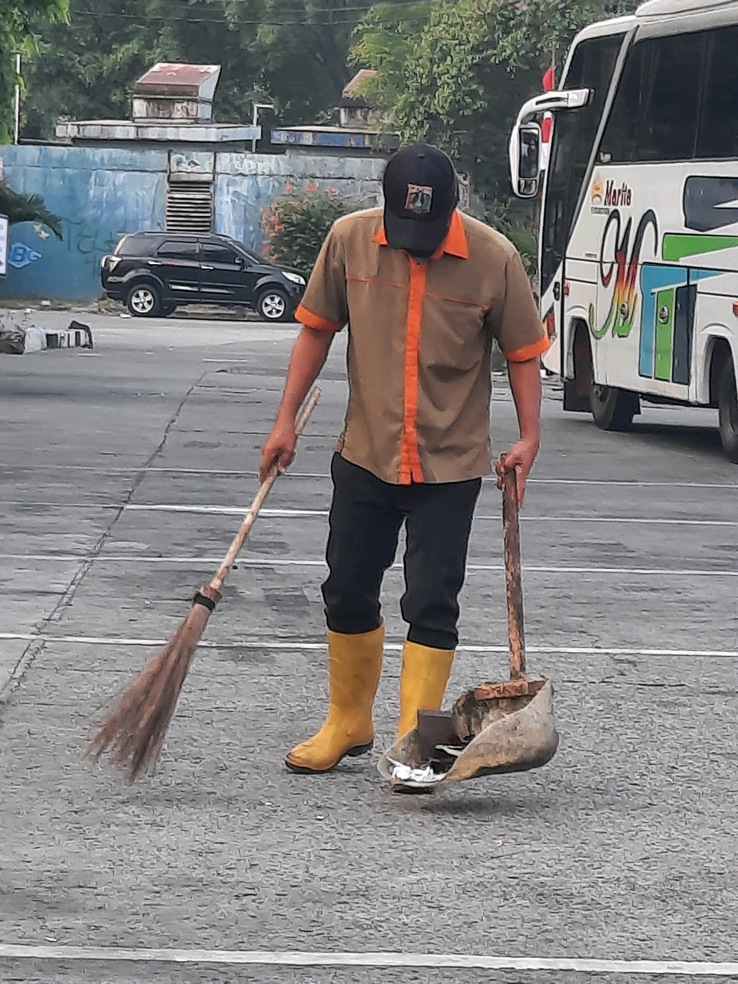 A man wearing a brown and orange uniform, black pants, and yellow boots is sweeping the ground with a broom. He is also holding a dustpan with a wooden handle, which appears to be made from a metal bucket or container. The man is standing in what looks like a parking lot or open paved area, with white lines marking parking spaces. In the background, there is a white bus with colorful graphics and the word "Marita" visible on its side. Further in the background, a black SUV is parked near a light blue wall. The scene appears to be outdoors, with trees and vegetation visible. This image was captured in East Jakarta, Indonesia.