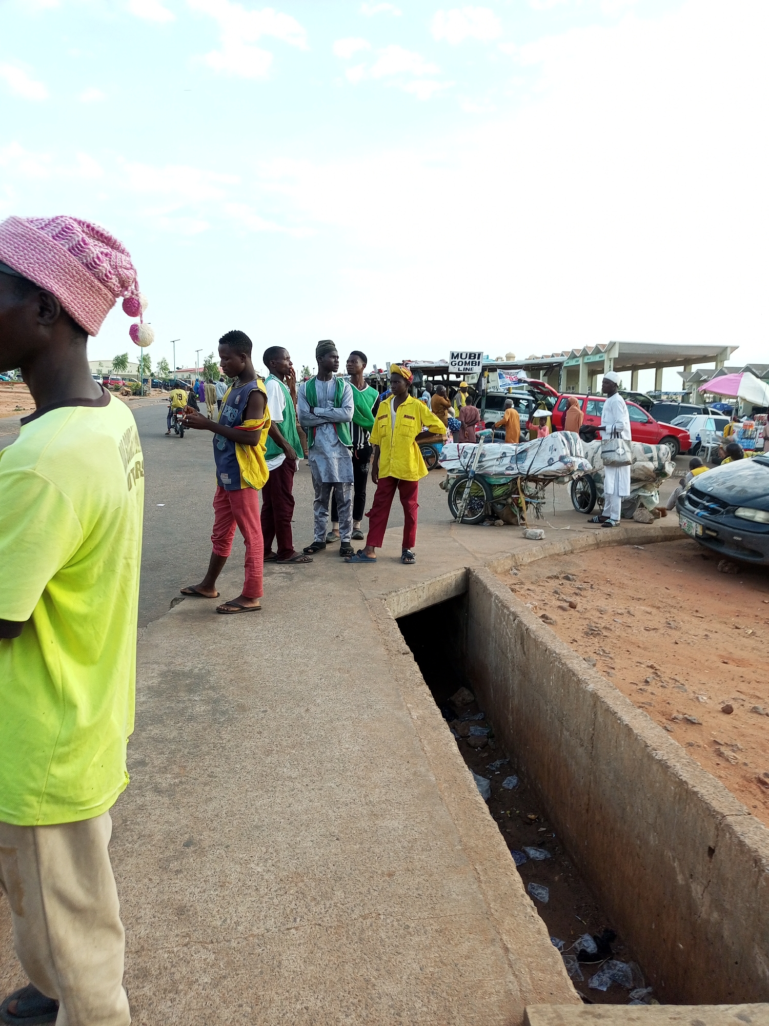 The image captures a street scene, likely a commercial area, with several people gathered along the side of a road. In the foreground, a person wearing a bright yellow t-shirt and light-colored trousers stands facing away from the camera, wearing a pink knitted hat with pom-poms. Behind them, a group of young men are standing on a concrete sidewalk. Some are in casual clothing, while others wear traditional attire like a green and white striped outfit and a gray agbada.
Further down the street, there's evidence of commercial activity. Several vehicles, including a red SUV, a black car, and a motorcycle loaded with goods, are visible. A sign can be partially seen that reads "MUBI GOMBI LINE," suggesting a transportation or commercial route. Stalls or makeshift shops are set up, with people attending to them. The environment appears to be dusty, with dirt roads and a concrete drainage ditch filled with debris in the lower right portion of the image. The sky is clear with a few scattered clouds. The overall impression is one of a busy, functional urban space.