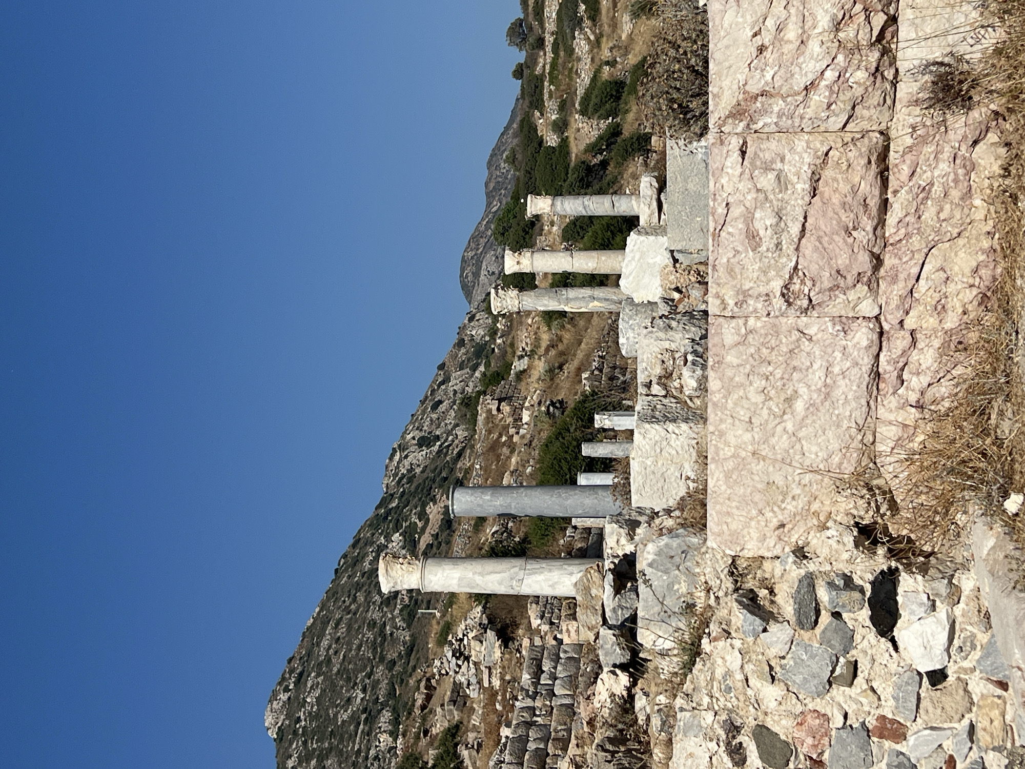 The image shows an ancient archaeological site with several weathered stone columns standing against a backdrop of a rocky, scrub-covered hillside under a clear blue sky. The columns vary in height and material; some are white marble with ornate capitals, while others are made of grey stone. In the foreground, there are large, rough-hewn stone blocks, some forming part of a wall. The site is littered with rubble and dry vegetation, indicating its age and exposure to the elements. The environment is arid and mountainous.