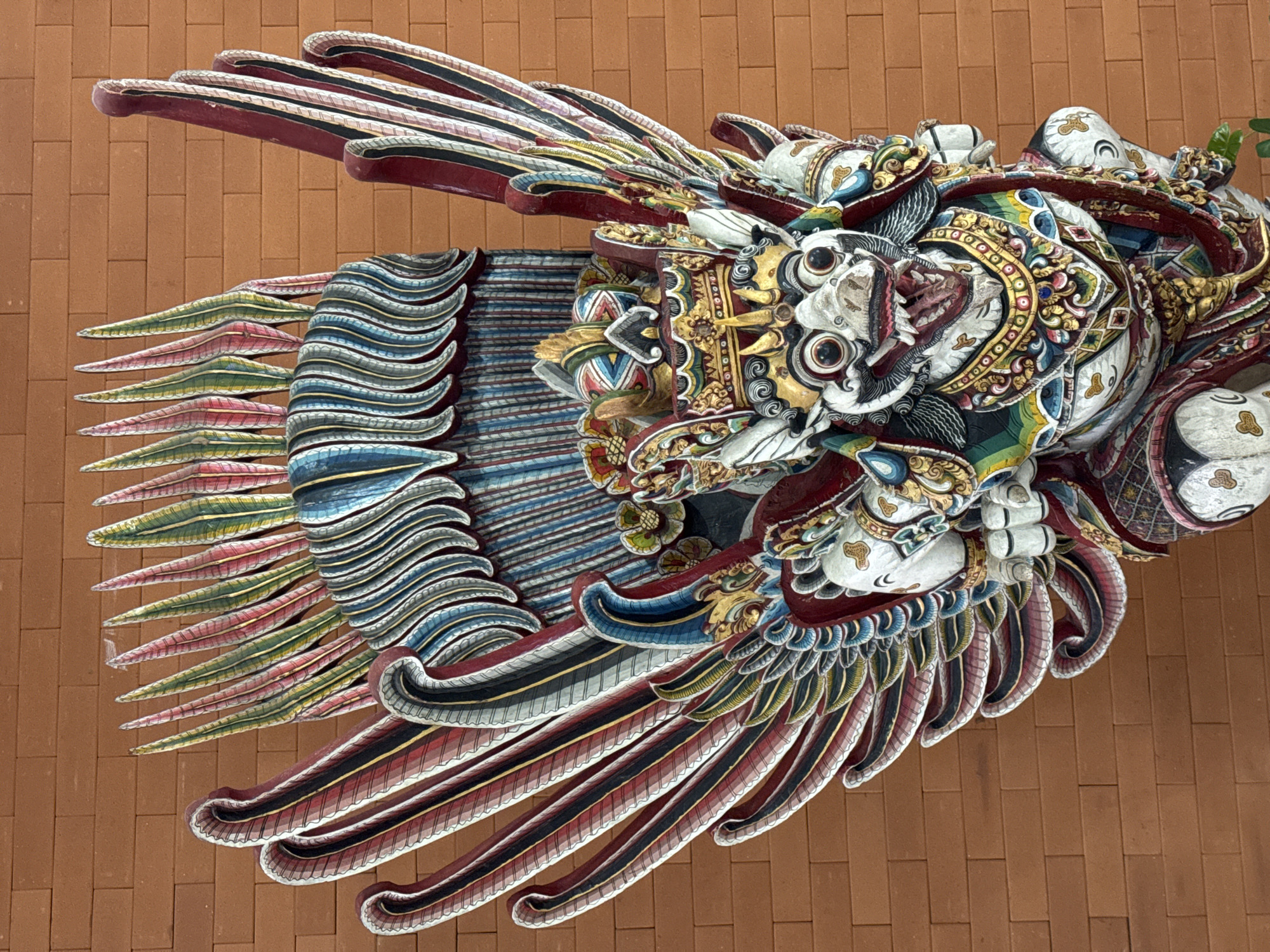 A detailed, colorful carved dragon adorns a wall. It is in the city of Unknown, Unknown. The dragon has multiple layers of intricate carvings. It has a fierce face with large eyes, an open mouth, and ornate head decorations. Its body is adorned with multicolored patterns and designs. A series of elaborate wings extend from its body, constructed from layered, curved, and feathered carvings. The background is a wall of brown bricks.