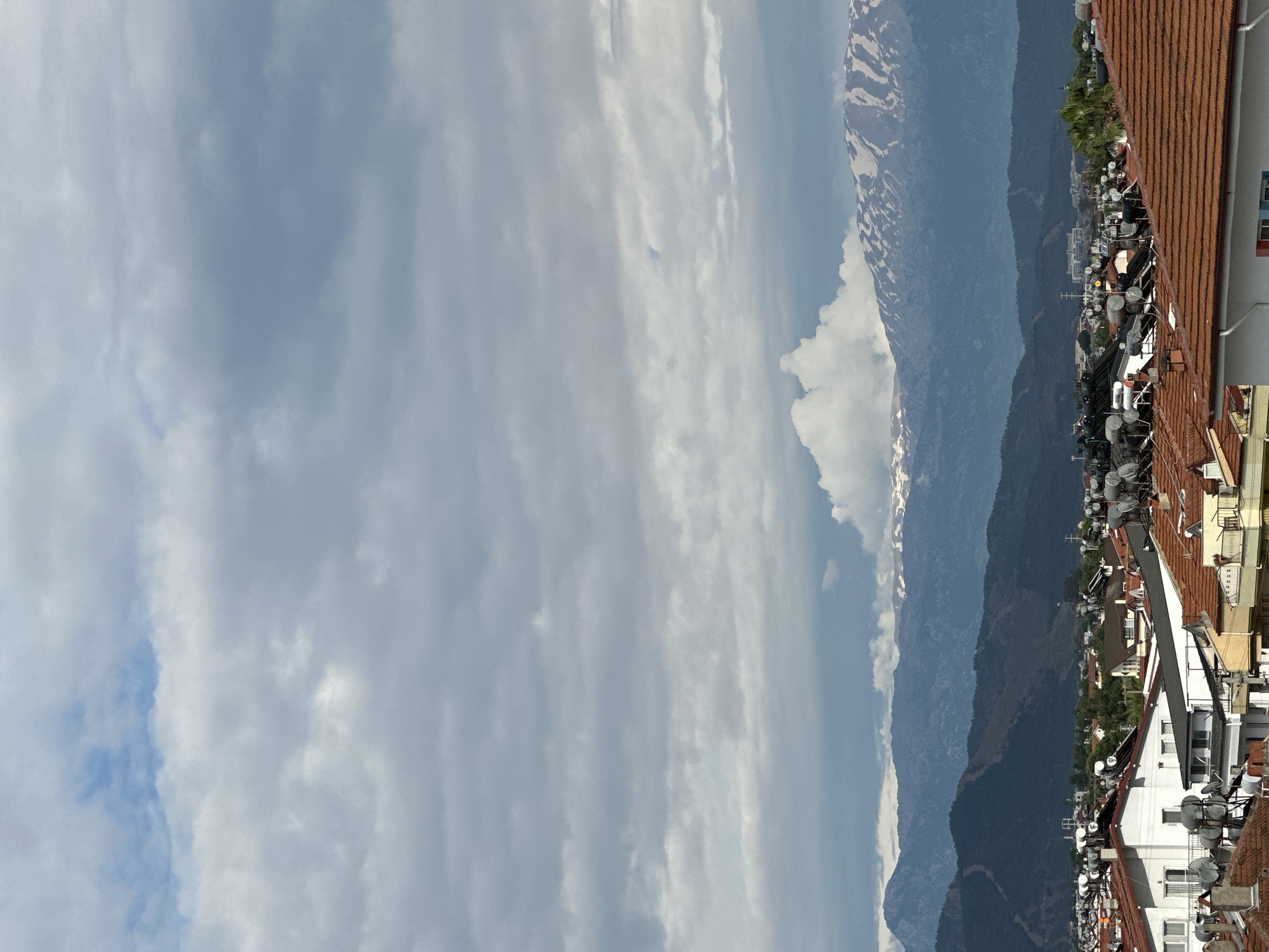 The image depicts a view from a building in Fethiye, Turkey, featuring a cityscape with mountains in the background under a cloudy sky. The foreground is dominated by the roofs of buildings, many equipped with satellite dishes. Mountains are visible in the midground with some snow at higher elevations. The sky is filled with a mix of light and dark clouds.