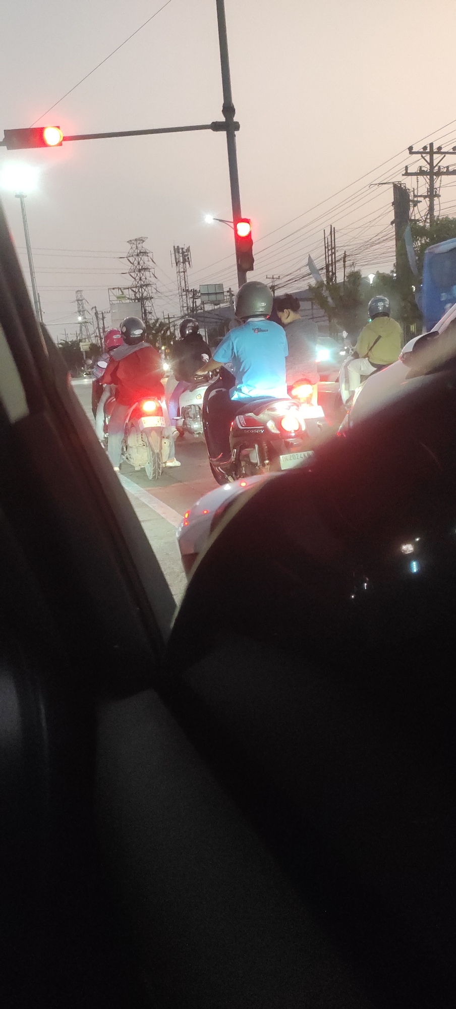 At dusk on a street in Jati Wetan, Indonesia, a line of motorcycles waits at a red traffic light. The sky is overcast, casting a soft, hazy glow over the scene. The foreground is dominated by the dark interior of a vehicle, with a portion of its windshield and dashboard visible. Beyond this, the street is filled with motorcyclists, many wearing helmets, their vehicles lined up in anticipation of the light changing. The traffic light itself is red, signaling a stop. In the background, power lines and towers suggest an urban or semi-urban environment. The overall impression is one of a typical traffic scene at the end of the day, with commuters navigating their way home.