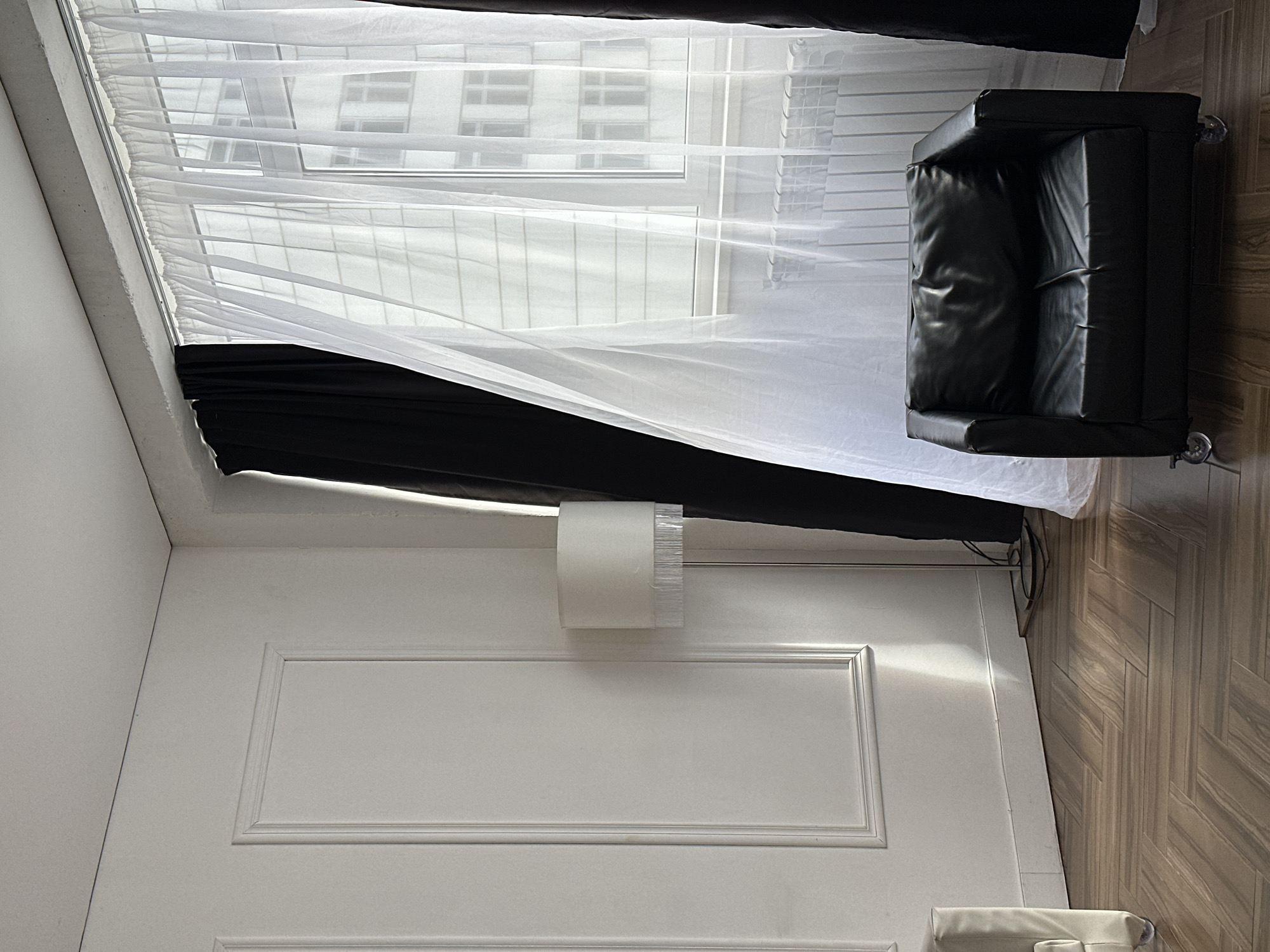 The image displays an interior room in Chaikovsky, Russia, featuring a large window with both sheer white and black blackout curtains. Bright natural light filters through the white curtains, revealing a view of a modern building with multiple windows outside.
To the right, a black leather armchair with a somewhat creased surface rests on a light brown wooden parquet floor, which exhibits a distinct herringbone or chevron pattern. The walls are painted white and feature decorative rectangular paneling. A tall floor lamp with a white cylindrical shade is positioned against the paneled wall.
The room is brightly illuminated, suggesting it is daytime. No people or specific activities are visible. The scene conveys a calm and organized atmosphere within what appears to be a modern residential space. There is no visible text.