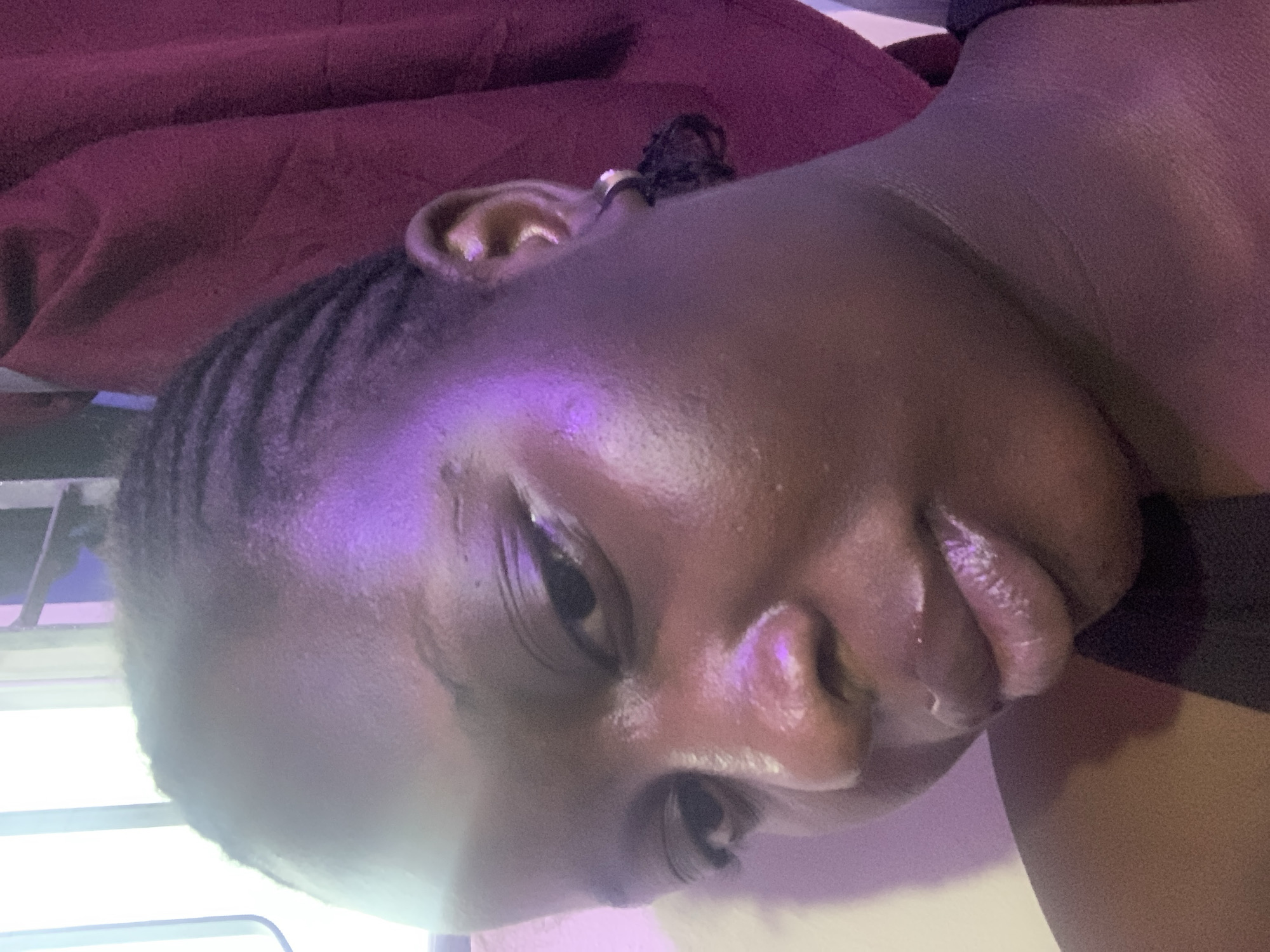 A close-up shot captures a person's face, angled from the right. Their dark skin has a dewy sheen, reflecting purple and white light. Braided hair is visible on the left side of their head, extending towards the back. The subject wears a black garment, with a burgundy fabric draped behind them. A small silver hoop earring with a black dangle is visible on their right ear. The lighting suggests an indoor setting, possibly at night. The location is not determinable from the image.