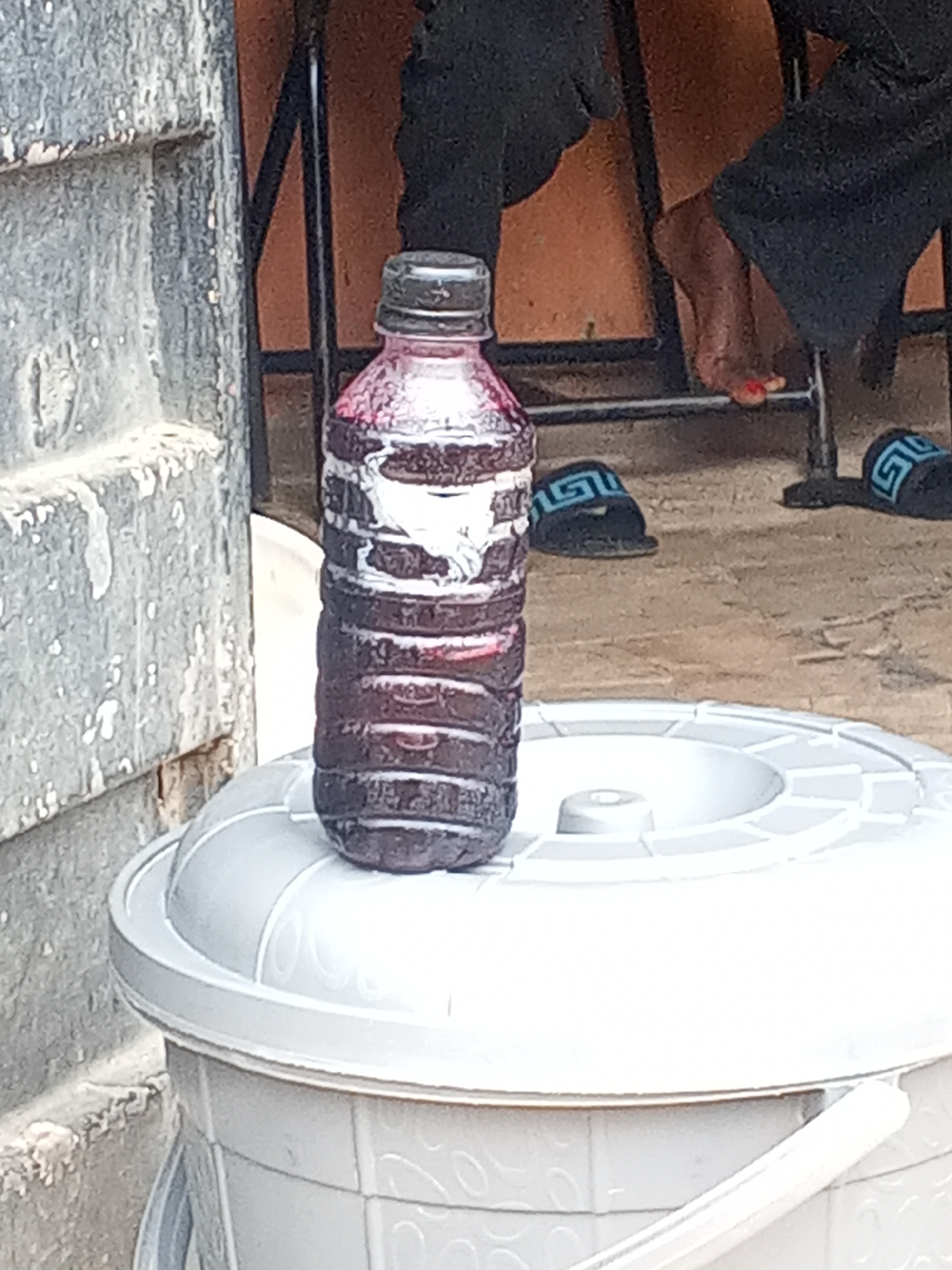 A plastic bottle containing a dark red liquid with white froth rests on top of a light grey bucket with a lid. The bottle is ribbed and has a dark cap. The bucket has decorative swirls on its side. To the left, there is a weathered grey doorframe. In the background, there's a metal chair with legs, occupied by a person wearing dark clothing. Their bare foot, with painted red toenails, is visible along with black sandals with blue designs nearby. The location is Gwong, Nigeria.