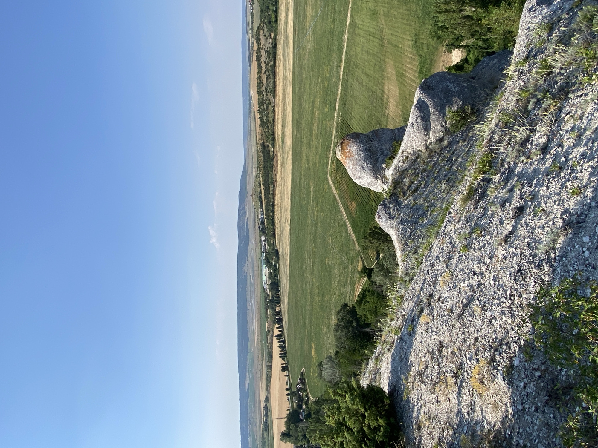 An elevated shot captures a vast landscape under a clear blue sky. In the foreground, rough, rocky terrain is covered with sparse vegetation. A large, weathered rock formation with a reddish-orange cap juts out on the right side of the foreground. Beyond this, a green field stretches out, with a distinct path cutting through it. Further in the distance, fields transition to a golden-brown hue, bordered by a line of trees. A small village or settlement is visible amongst the trees and fields, with buildings and roads. The horizon is marked by rolling hills and more distant, hazy mountains. The overall scene depicts a rural, agricultural landscape bathed in daylight.