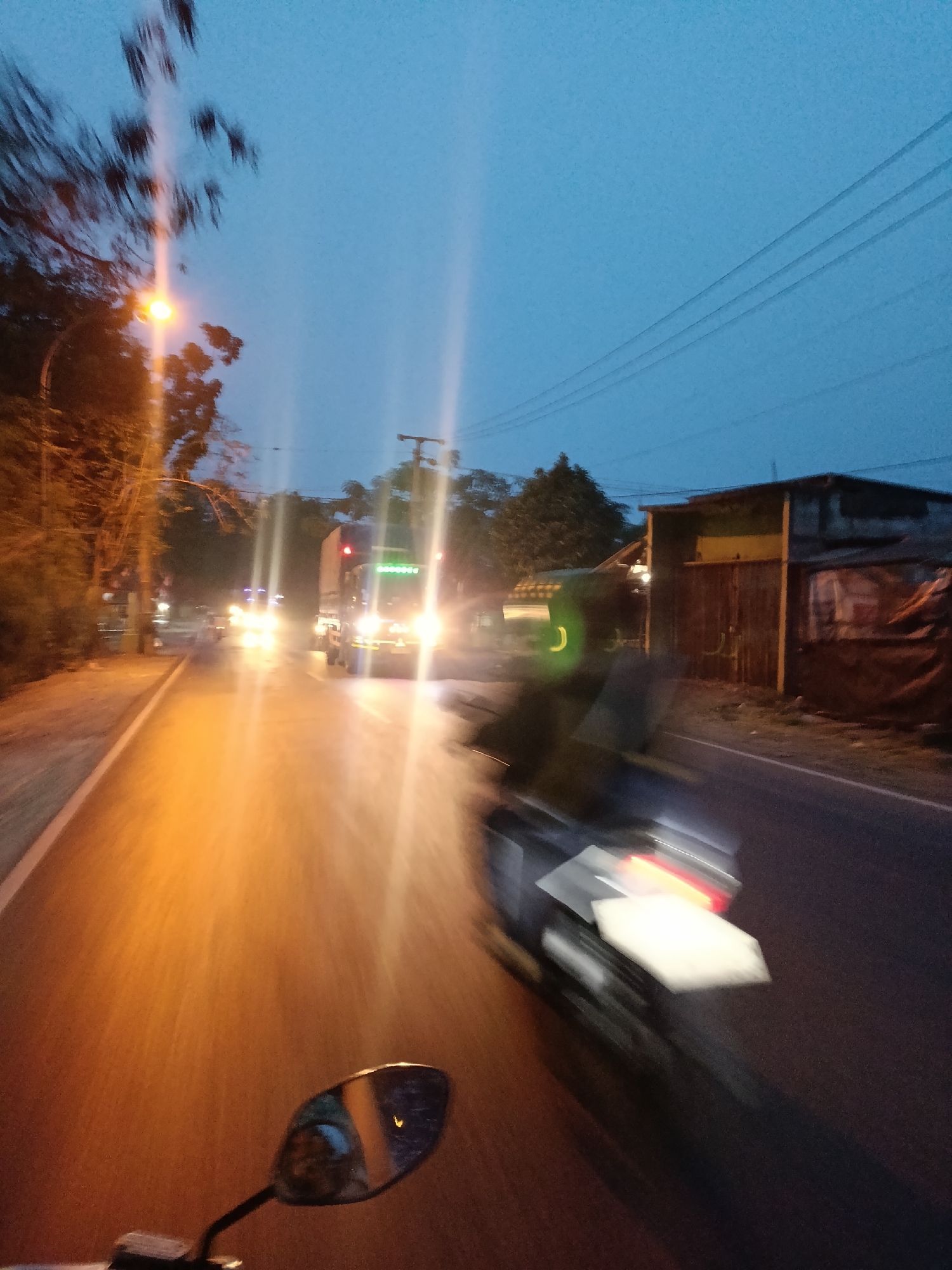 The image depicts a blurry, motion-filled scene at dusk on a road in Jonggol, Indonesia. The perspective is from a moving vehicle, likely a motorcycle given the presence of a rearview mirror in the foreground. A streetlight with an orange glow illuminates the left side of the road, casting light on trees and foliage. Further down the road, headlights of oncoming vehicles, including trucks and other cars, create streaks of light due to the motion blur. The right side of the road features a building with a weathered facade, possibly a shop or dwelling, with some tarpaulin covering. Power lines are visible against the darkening blue sky. A distinct blur of a motorcycle is visible in the middle ground, suggesting the photographer is riding alongside or behind it. The overall impression is a busy road at twilight in a semi-urban or rural setting.