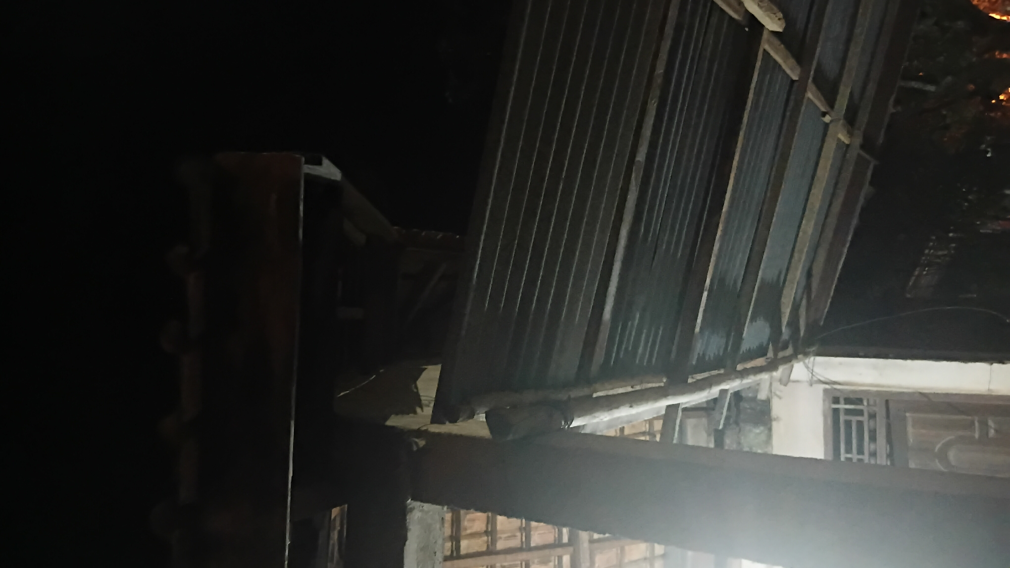 A corrugated metal roof is tilted precariously on a wooden support structure. The roof appears damaged and is made of overlapping metal sheets. The scene is dark, suggesting it is nighttime or indoors with limited lighting. The background is indistinct and mostly black, but hints of a building with a wooden door and window are visible. There are no people or animals in the image. The location is identified as Prabumulih, Indonesia.