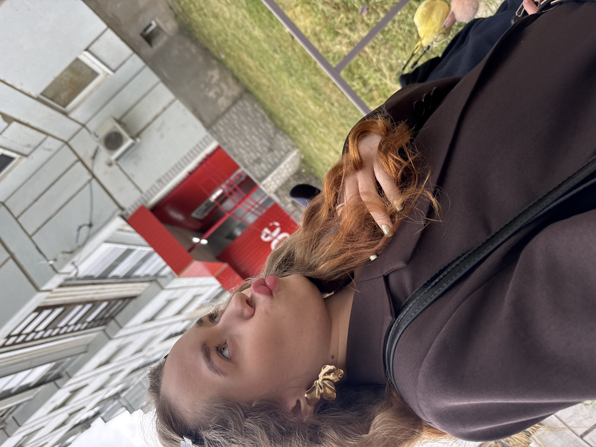 A young woman, likely in her late teens or early twenties, is the central subject of this image, oriented vertically (rotated 90 degrees counter-clockwise). She has long, reddish-brown hair with lighter streaks and is wearing a dark brown jacket or coat with a black strap visible over her shoulder. A large, gold-colored, sculptural earring adorns her visible ear. Her fingernails are painted a light green or iridescent color. She is posing with her lips slightly pursed, looking upwards and to her left, suggesting a stylized expression for a photograph.
The background depicts an urban or residential outdoor setting, consistent with Chaikovsky, Russia. To the left, a multi-story building with light-colored panels and multiple windows, along with an external air conditioning unit, is visible. Behind the woman, a prominent red architectural feature, possibly an entrance or a facade element, stands out. Partial white text, appearing to be "Э0" or "30," is visible on this red structure. Further in the background, green grass, a paved area, and a light-colored metal railing are present. The scene is illuminated by daylight, likely under an overcast sky, as there are no harsh shadows.