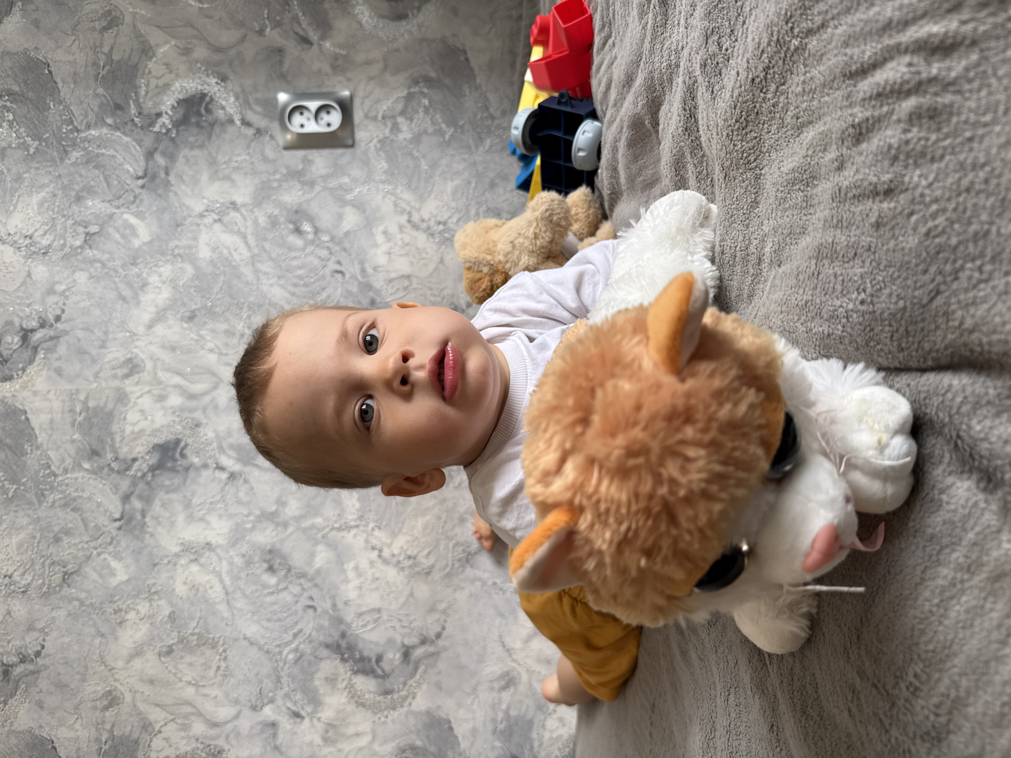 A baby is lying on a gray blanket next to a stuffed toy cat. The location is Rusanovo, Russia. Toys are in the background, near a wall with a light switch.