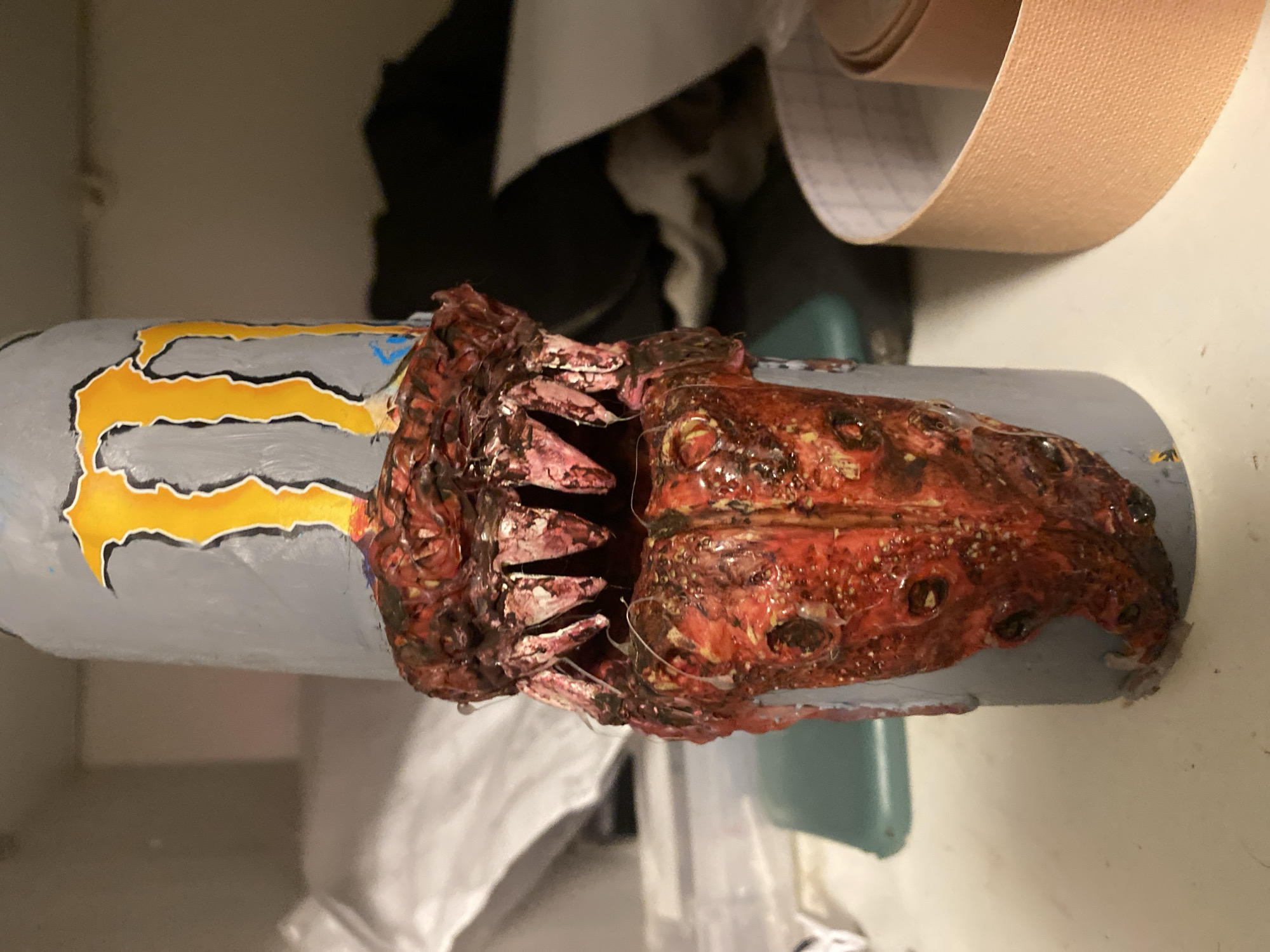 A Monster Energy drink can has been modified to have an artificial monster mouth affixed to it. The can is painted gray with the yellow Monster logo on the left side. The artificial mouth is red and brown, with visible teeth. The background includes a roll of tape, a white piece of cloth, and a teal object. The scene is indoors. There is no indication of the location being Ternopil, Ukraine, or any other city. There are no people in the image.