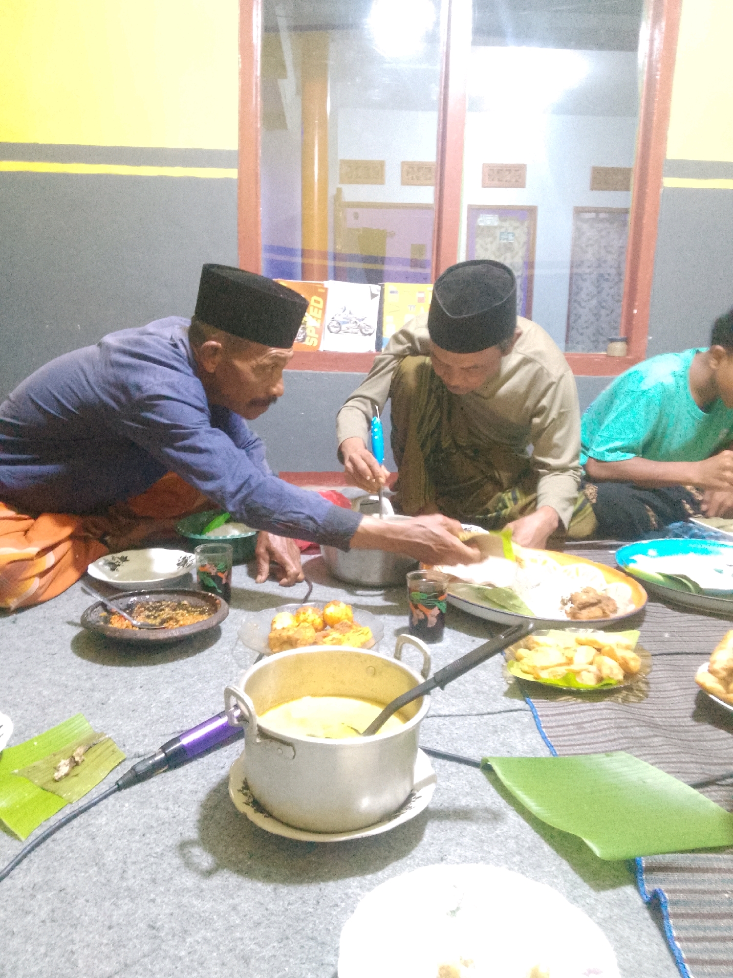 In Krucil, Indonesia, a group of men are gathered on the floor for a meal. One man on the left, wearing a blue shirt and a black cap, is seated with his legs tucked. Another man in the center, in a tan shirt and black cap, is also seated and appears to be serving food from a pot. A third man, in a teal shirt, is partially visible on the right, also seated. The floor is covered with a gray carpet, and various dishes of food, including rice, vegetables, and fried items, are spread out on plates and banana leaves. A microphone is placed near a pot of soup. The background features a window with a view of a building and some wall decorations, suggesting an indoor setting.