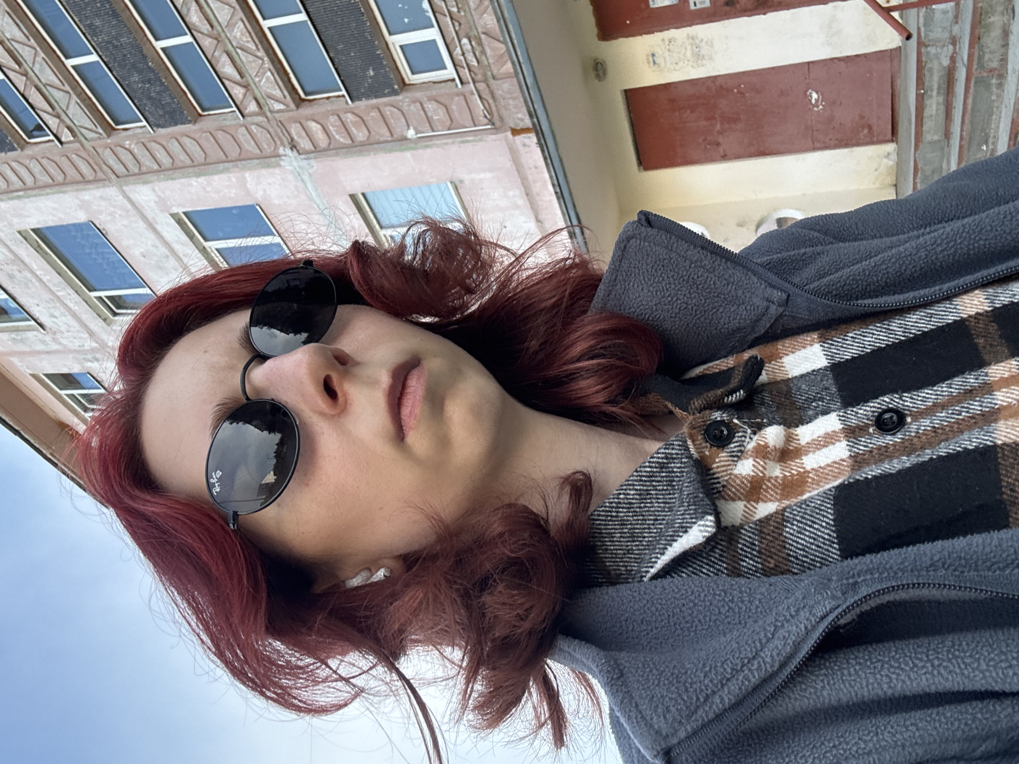 In a medium shot, a person with striking red hair, wearing sunglasses and a plaid shirt under a grey jacket, looks directly at the camera. The person is outdoors, with a building of apartment-style housing in the background. The building has multiple floors with windows and balconies. The sky is visible above. The ground in front of the building appears to be paved with stairs leading up to a doorway. The overall scene suggests an urban environment.