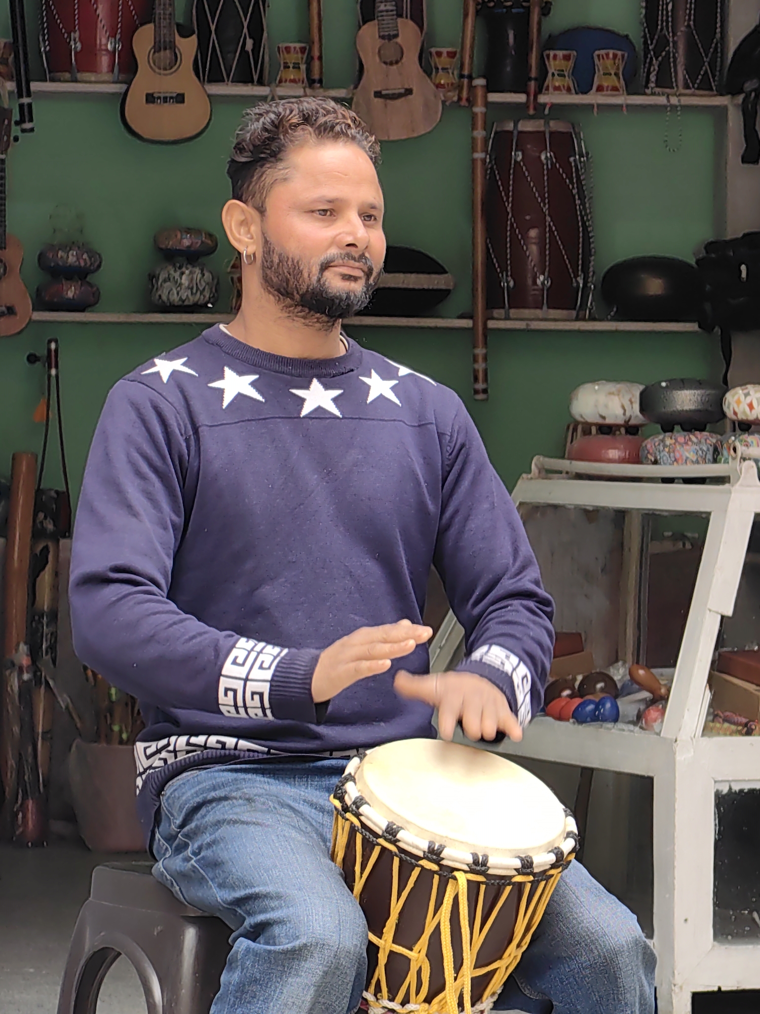 In Vashisht, India, a man with a beard and a hoop earring plays a djembe drum. He is seated on a black stool and wears a navy blue sweatshirt with white stars and a white geometric pattern around the cuffs and waist. He is also wearing blue denim jeans. His hands are positioned over the drum, with his left hand in mid-air and his right hand in contact with the drumhead, indicating he is actively playing. The background shows shelves filled with various musical instruments, including guitars and other types of drums. The setting appears to be a shop or workshop specializing in musical instruments.