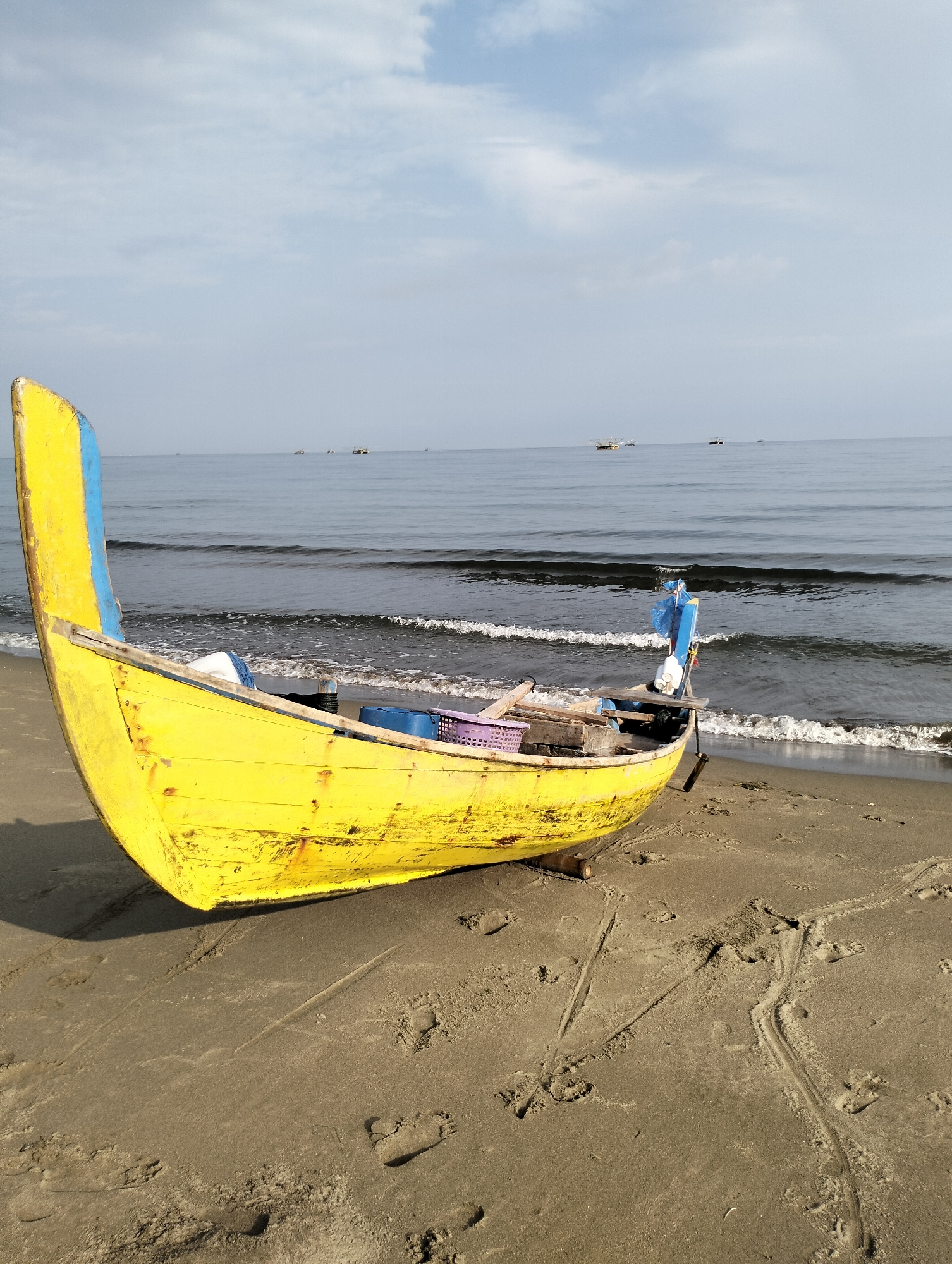 A yellow and blue wooden boat sits on a sandy beach at Curehtunong, Indonesia. The boat is angled towards the viewer, with its bow on the left side of the frame. There are objects inside the boat, including a purple basket. The beach shows numerous footprints and drag marks in the sand. In the background, the ocean is calm with small waves lapping at the shore. Several distant boats are visible on the horizon. The sky is overcast with patches of blue.