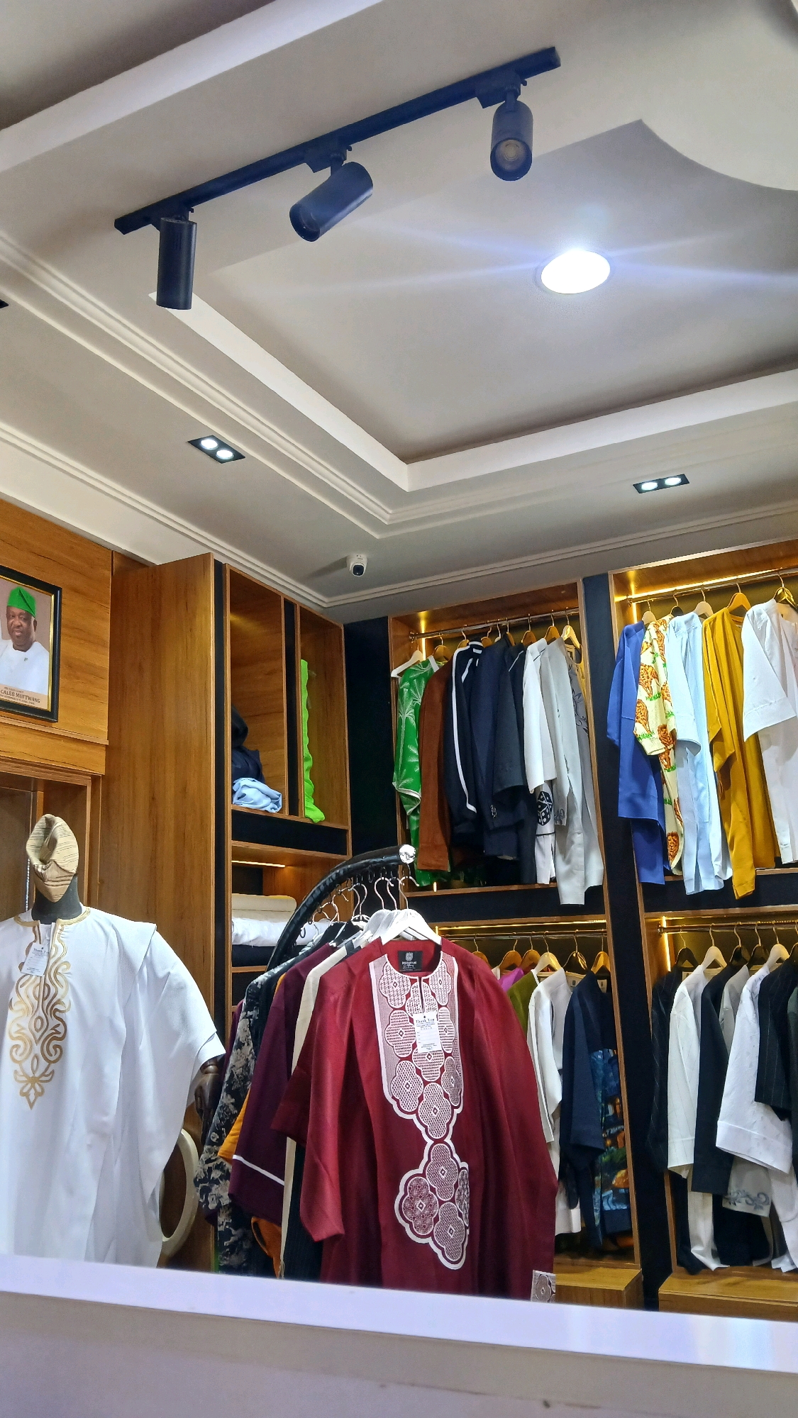 The image shows the interior of a clothing store in Gura Top, Nigeria. The store features an overhead track lighting system with multiple adjustable spotlights. The walls and shelves are made of light-colored wood. Various garments, including traditional African attire, are displayed on hangers. In the foreground, a mannequin is dressed in a white kaftan with gold embroidery. A framed portrait of a man wearing a green cap is visible on the left wall. The scene is well-lit and organized, suggesting a retail environment.