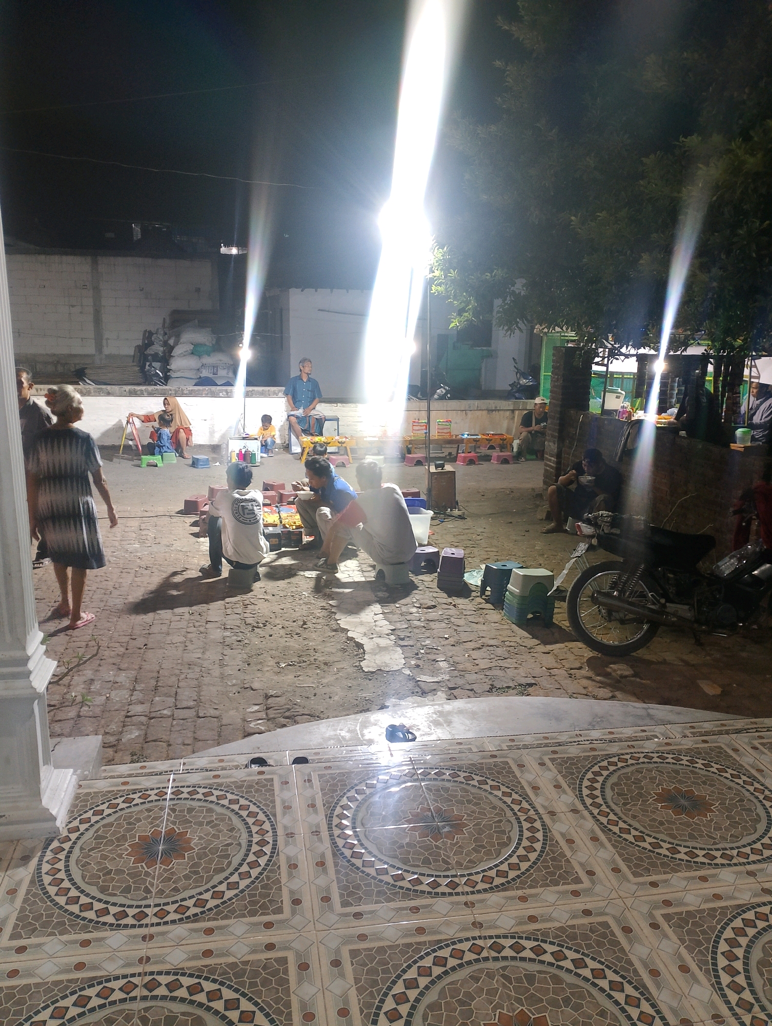 This is a nighttime outdoor scene, likely in Kedungsari, Indonesia, with a focus on a gathering of people. The foreground is dominated by decorative patterned tiles. Beyond the tiles, a brick-paved area is visible, where several individuals are seated and interacting.
In the mid-ground, a woman in a patterned dress walks away from the camera. Nearby, two men sit on the ground, one facing away from the camera, the other facing right and appearing to eat. A motorcycle is parked to the right of these men. Further back, several other people are seated on low stools or the ground, some appearing to be engaged in conversation or eating. One man in a blue shirt is seated on a raised platform on the far right. In the background, there are buildings and stacked sacks, illuminated by bright lights that create lens flares in the image. The overall atmosphere suggests a casual gathering or market setting, possibly a street food stall or local event.
