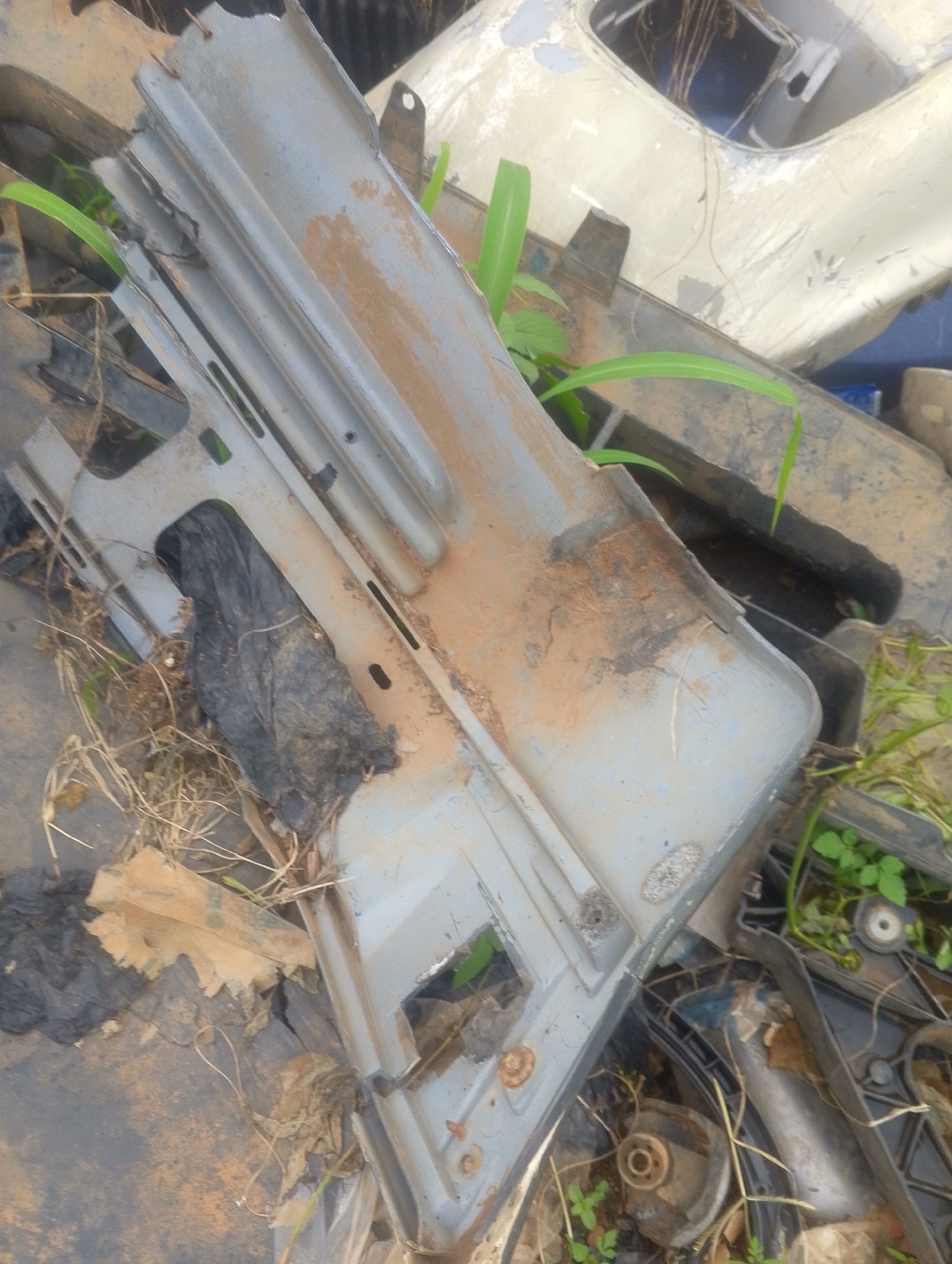 The image depicts a pile of discarded vehicle parts, possibly from a junkyard or scrap area, located in the city of Jos, Nigeria. The central object is a large, grey, metallic piece, likely a fender or bumper, showing signs of rust and dirt. Several green plants are growing amidst the debris, indicating an outdoor environment. Black plastic bags and dry grass are also scattered around the area. The overall scene suggests an accumulation of waste materials.