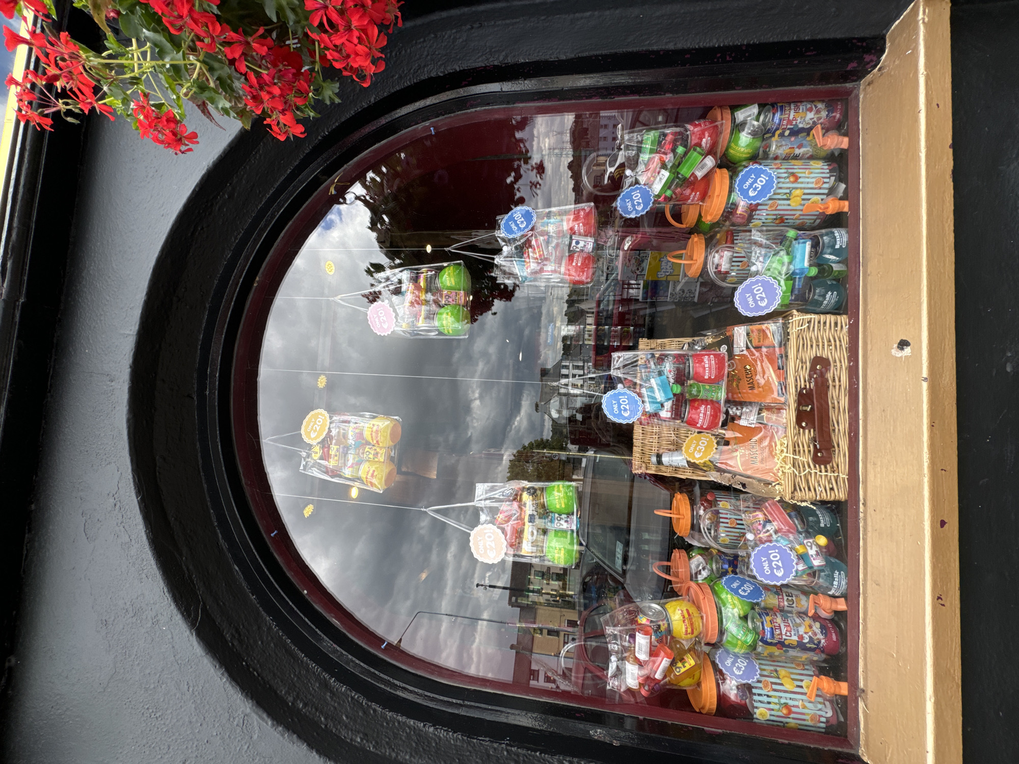 This image captures a shop window display in Waterford, Ireland, featuring an assortment of colorful candy bags and jars filled with sweets. The window is framed by a dark, arched structure. Hanging from the top of the window are several clear plastic bags, each containing various candies and marked with price tags reading "ONLY €20!".
Below the hanging bags, a woven basket sits prominently, holding more packaged sweets and bottles. To the left of the basket are large clear jars filled with candies, some with orange lids and taps. These jars are also adorned with price tags, indicating "ONLY €30!". Several other clear bags and containers of candy are visible throughout the window, some labeled with "ONLY €20!". The display is vibrant and appears to be a sweet shop or a similar establishment. Reflections of the cloudy sky and buildings across the street are visible on the window glass. Red flowers hang from the top right corner of the frame, adding a touch of natural color to the scene.