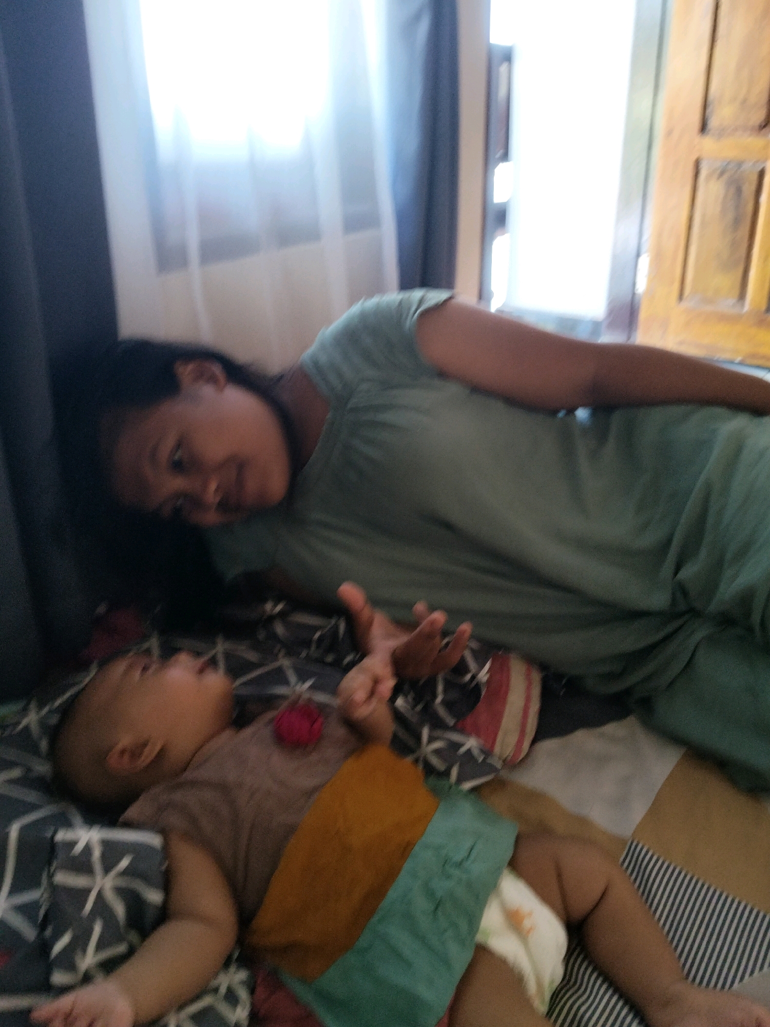 A mother is lying down next to her baby on a bed in a room in Gempolan, Indonesia. The mother, wearing a green top, is looking at her baby. The baby, wearing a brown shirt and a diaper, is lying on its back. The room has a window with sheer white curtains and a dark blue curtain on the left side. A wooden door is visible on the right side of the image. The bed is covered with a patterned blanket.