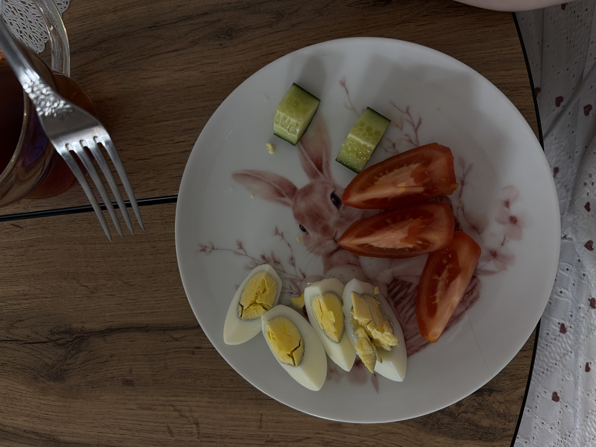 An overhead shot captures a simple meal prepared and laid out on a dark brown wooden table, indicative of an indoor setting, potentially in Reshetnikovo, Russia. The main subject is a white, round plate featuring a charming printed design of a rabbit and subtle pinkish-brown floral or branch patterns.

Arranged on the plate are several food items: four halves of hard-boiled eggs, showing their yellow yolks (one piece appears slightly broken), four wedges of bright red tomato, and two rectangular chunks of green cucumber. To the upper left, a clear glass or mug containing a dark reddish-brown liquid, likely tea, is partially visible. A silver fork with decorative detailing on its handle rests beside the glass, extending towards the plate. The wooden table surface exhibits a distinct grain. To the right side of the plate, a section of white fabric with a patterned, possibly embroidered or lacy, texture can be seen. The scene suggests a ready-to-eat breakfast or a light snack. No text is visible in the image.