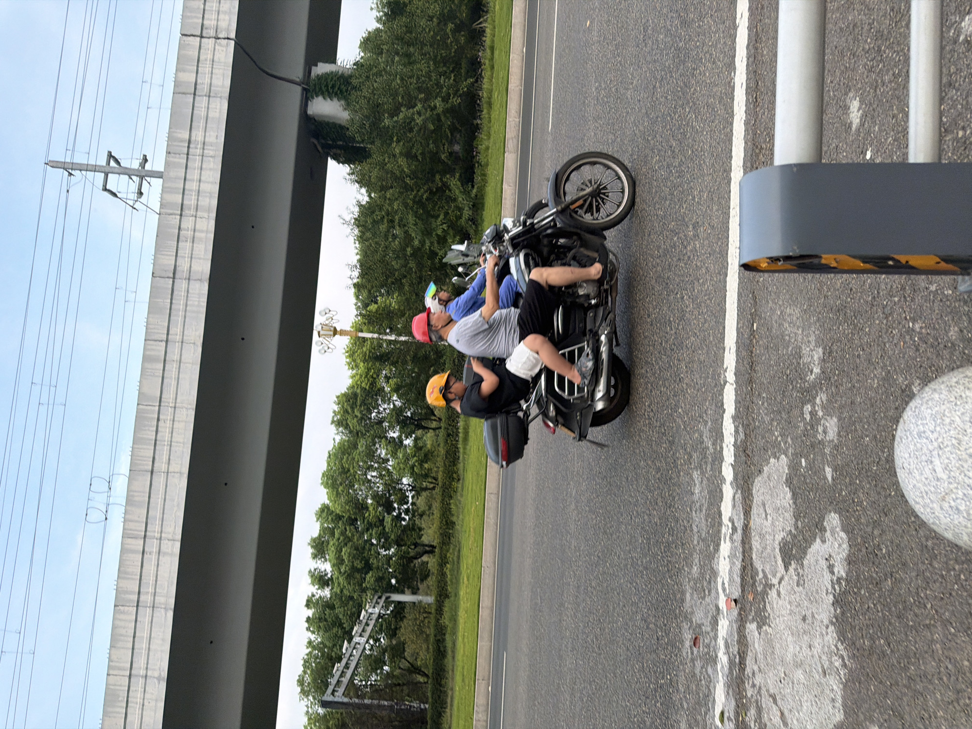 Three individuals, two adults and one child, are riding on a motorcycle on a paved road in Jiangjin District, Chongqing, China. The adult in the front is wearing a red helmet and a grey t-shirt, with black shorts and a white bag. The adult behind him is wearing a blue outfit. The child, positioned at the rear of the motorcycle, is wearing a yellow helmet and a black t-shirt with white shorts. The motorcycle has a black storage box mounted behind the seating area. They are traveling alongside a grassy area and trees, beneath a large elevated concrete structure, possibly a highway overpass, with visible overhead power lines. The road surface shows some wear and markings, including a white line. In the foreground, a grey metal barrier with yellow and black hazard stripes is partially visible. The sky is overcast.