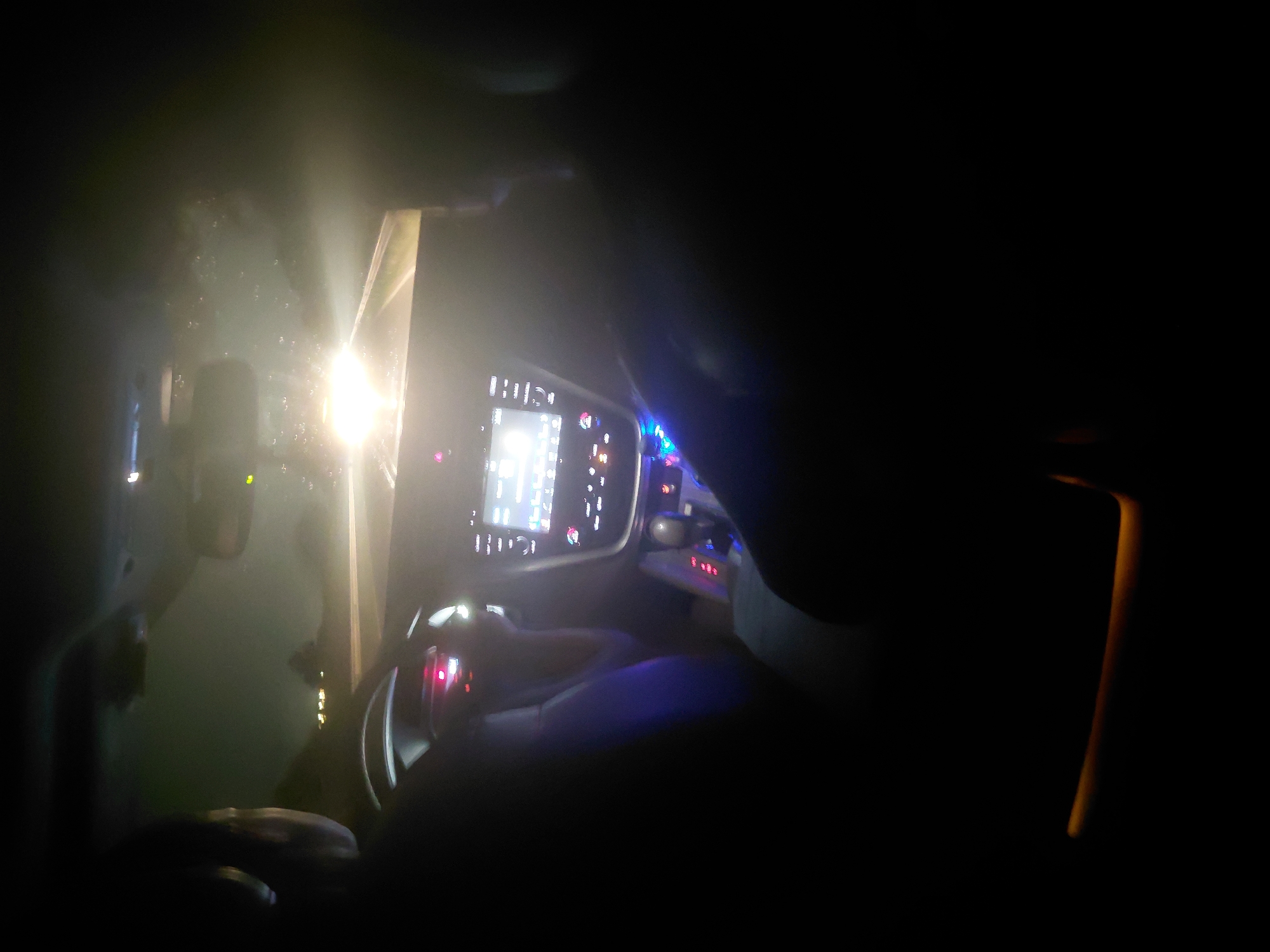The image depicts the interior of an automobile at night, viewed from a rear passenger perspective in Radinovo, Bulgaria. A driver is partially visible on the left, positioned behind a steering wheel. A front passenger is also partially visible on the right. The vehicle's dashboard features a central, illuminated digital infotainment screen displaying a graphical user interface. Below it, the center console contains a gear selector and multiple backlit control buttons, illuminated with red and blue light. The windshield is visibly wet, presenting numerous water droplets or smudges, and reflects an extremely bright, centralized light source from outside the vehicle, likely oncoming headlights, which obscures the view forward. Less intense, distant lights are also visible through the upper section of the wet windshield. The overall scene is dark, with internal illumination primarily from the vehicle's controls and screen, complemented by a subtle amber light source visible in the lower right foreground of the cabin, possibly ambient lighting. The vehicle appears to be in motion.