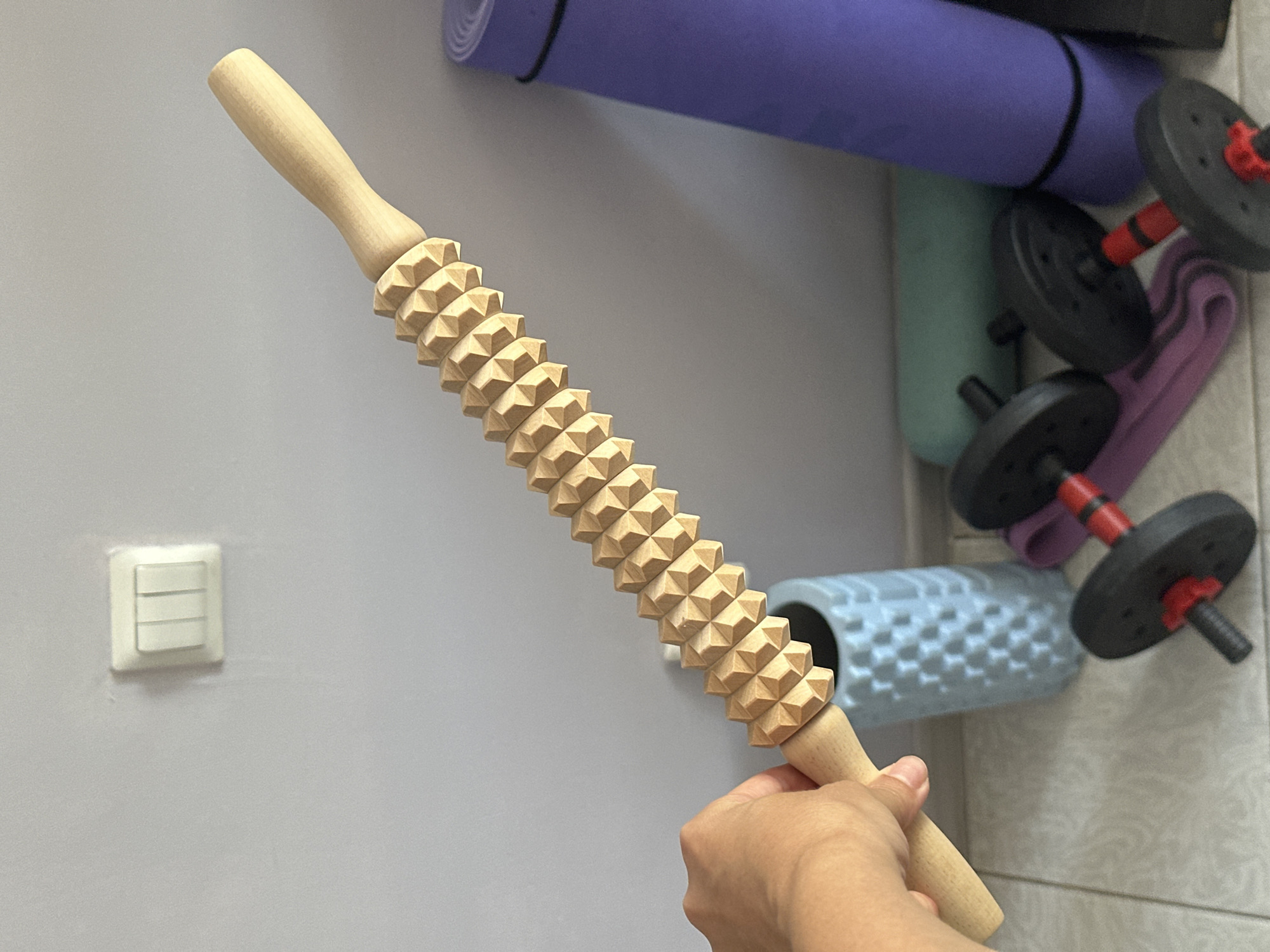 A wooden massage roller with a textured surface is held by a hand against a pale grey wall. To the right of the roller are two rolled-up yoga mats, one purple and one green, and a light blue textured cylindrical object. In the foreground, two dumbbells with black weights and red accents are placed on the floor, along with a purple resistance band. A white light switch is visible on the wall behind the roller. The scene appears to be indoors, possibly in a home or a gym. There is no discernible information about the city of Sochi, Russia in the image.