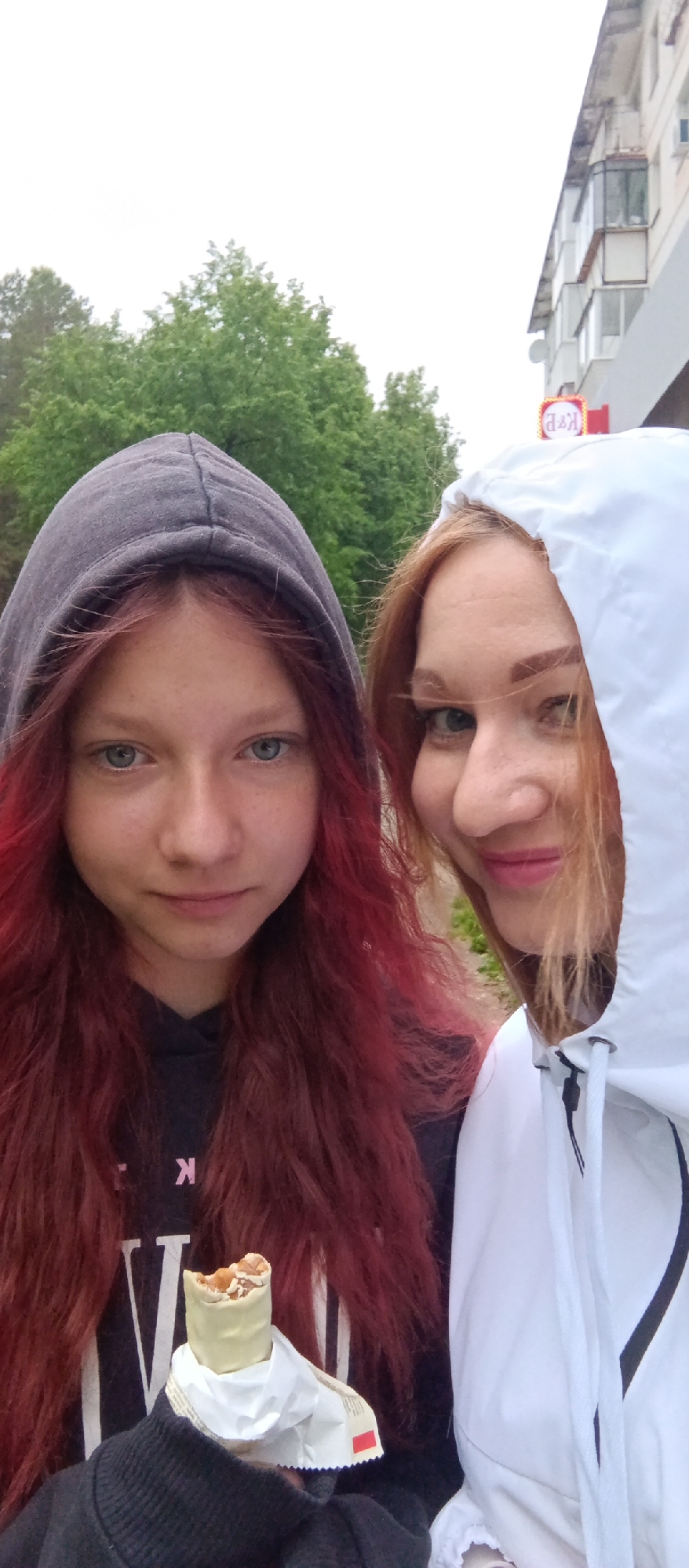 Two females are standing outdoors in what appears to be an urban environment.
The female on the left has red hair and blue eyes, and she is wearing a dark-colored hoodie. She is holding a white-covered snack in its packaging, partially eaten.
The female on the right has reddish-blonde hair and is wearing a white hooded jacket.
In the background are trees and a building. A sign with "K&E" is visible.