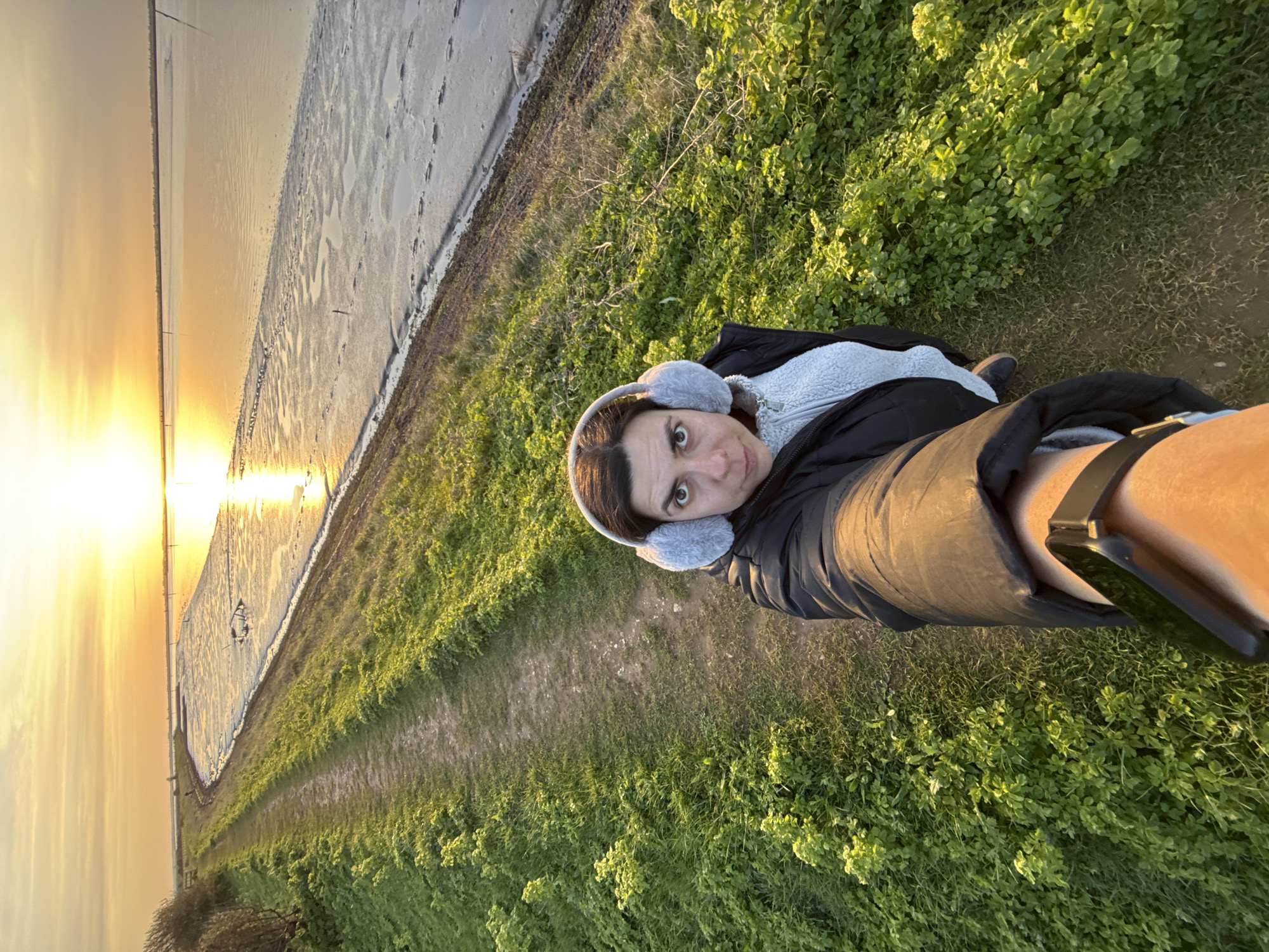 A medium-shot, point-of-view photograph shows a woman in ear muffs posing in a meadow at sunset, possibly in Great Yarmouth, UK. She wears a dark coat over a white jacket. The sun reflects across a body of water in the background. A path of grass is next to her. Her outstretched arm holds a smartphone, with the band of a smartwatch also visible.