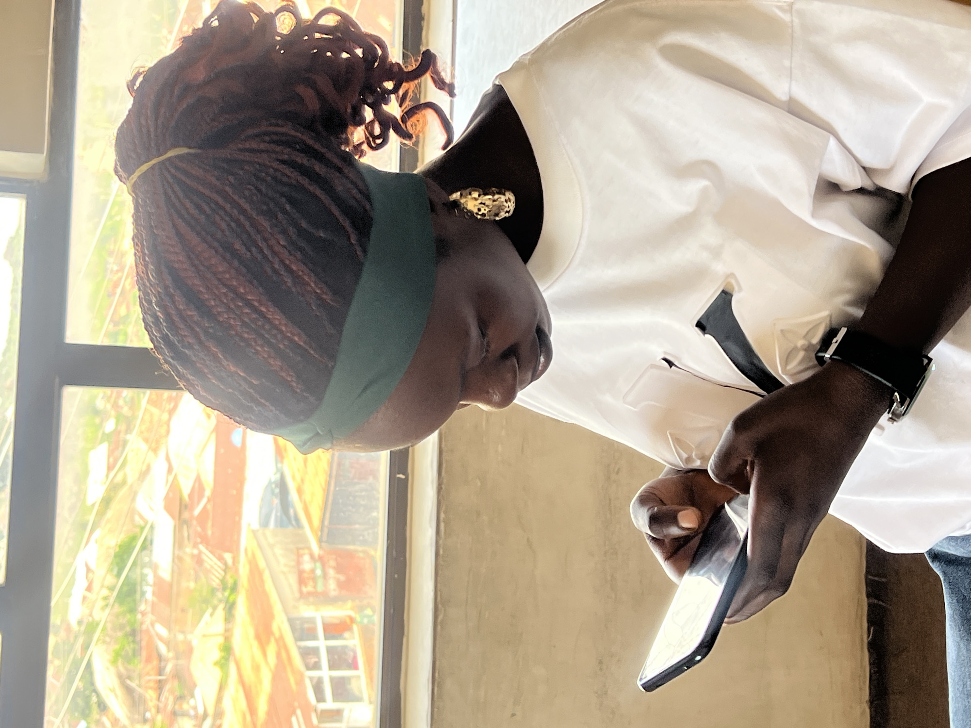 A medium shot captures a young Black woman with braided hair pulled back in a ponytail, wearing a green headband and a white t-shirt with a black design, looking down at her phone. She is seated indoors, possibly in a room with a large window that offers a view of a densely populated urban area with corrugated metal roofs, indicative of a city like Jos, Nigeria. The woman's hands are holding the phone as she interacts with it. A gold earring dangles from her left ear, and a black watch is visible on her left wrist. The lighting suggests it is daytime, with sunlight streaming in from the window, creating bright highlights and some lens flare. The background outside the window shows a typical cityscape with residential buildings and vegetation.