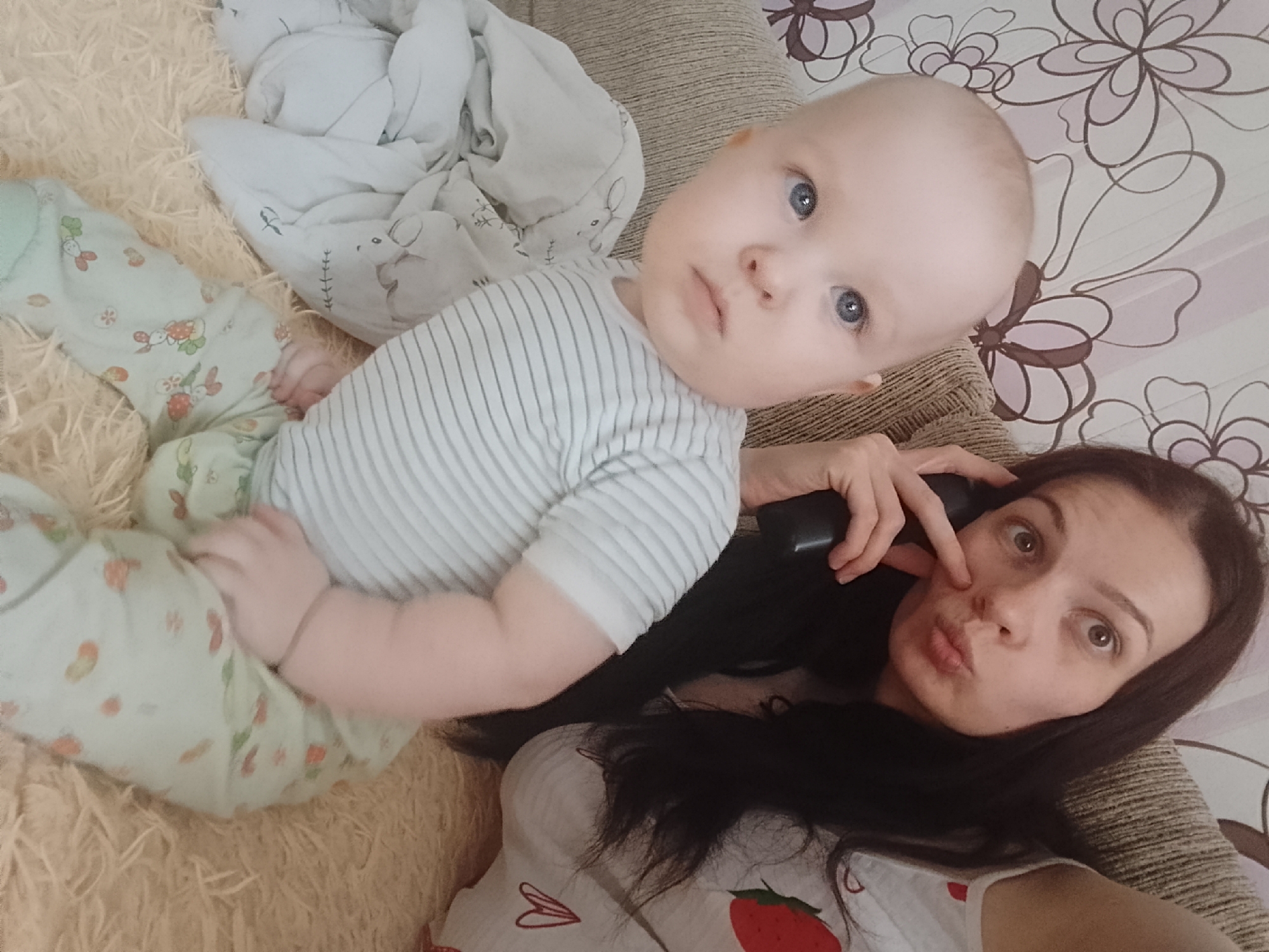 This is a close-up, over-the-shoulder selfie taken in an indoor setting, likely a home environment. A woman with dark, long hair is in the foreground, holding a dark object, possibly a phone, to her ear and pouting her lips. Her eyes are wide open, looking at the camera. To her left and slightly behind her is a baby with blue eyes, wearing a striped shirt and a pastel green onesie with a strawberry and flower print. The baby is lying down, with its hand resting on its stomach. The background consists of a beige, textured surface and a cream-colored blanket or sheet with a floral pattern. The overall lighting suggests a well-lit room, possibly with natural light. There are no discernible indicators of the specific location within the city of Minsk, Belarus.