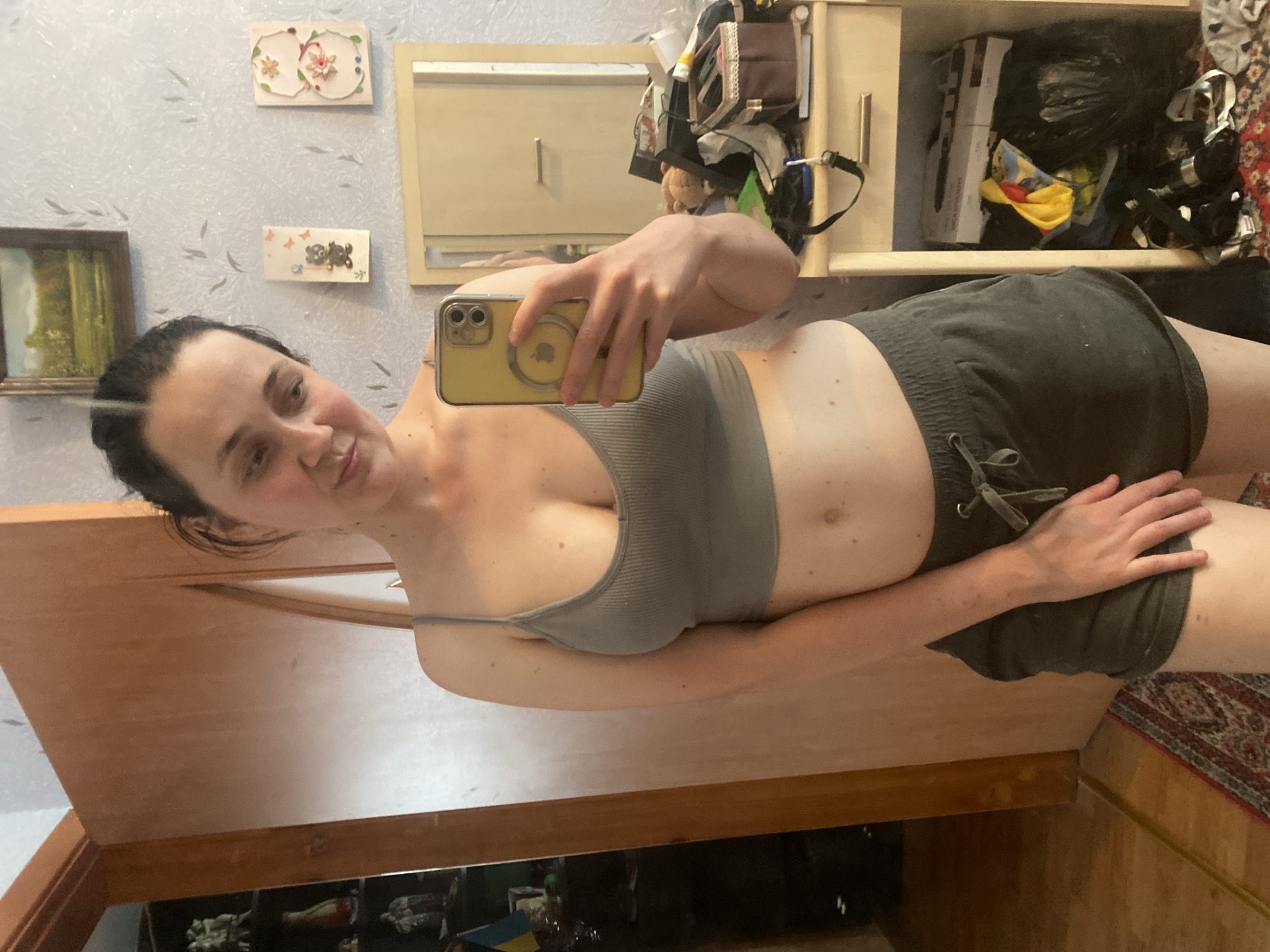 A young woman, with dark hair pulled back, poses for a mirror selfie in what appears to be a bedroom. She is wearing a grey sports bra and dark grey shorts. She is holding a yellow iPhone in her right hand, angled to capture her reflection. Behind her, a closet door is visible on the left, and a dresser with a mirror is on the right. The dresser top is cluttered with various items, and shelves below are filled with boxes and bags. A painting hangs on the wall above the dresser, along with two small decorative pieces. A rug is visible on the floor in the foreground. The overall impression is of a casual, domestic setting.