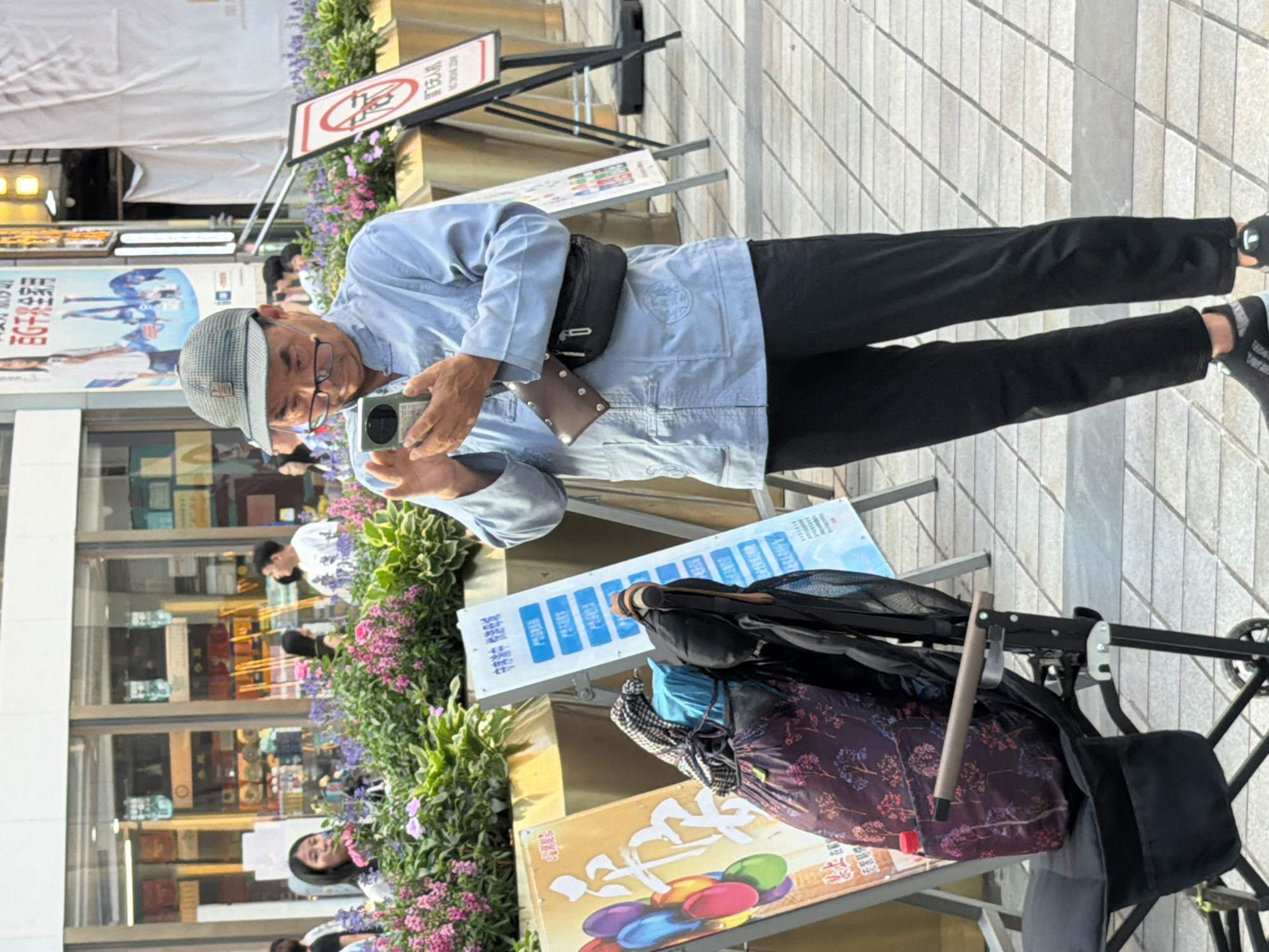 In Yuzhong District, China, an elderly Asian man wearing a grey flat cap, glasses, and a light blue long-sleeved shirt stands on a tiled pedestrian walkway, holding a mobile phone in his hands as if taking a picture or video. He is wearing black pants and black athletic shoes with white accents. A dark brown wallet-like pouch is clipped to his shirt, and a black fanny pack is worn around his waist. Behind him, to his left, is a child's stroller with a patterned bag and a blue covering draped over it, along with a partially visible red water bottle. Two signs are positioned behind the stroller; one on the left has a colorful balloon graphic and Chinese text, while the other, on the right, is white with a red circular sign with a diagonal line, indicating prohibition, and Chinese text below. The man is facing slightly to his right and looking down at his phone. In the background, there are businesses with glass storefronts and displays, some with posters featuring people. There are also planters filled with pink and purple flowers and green foliage along the street. Other people are visible in the background, including a young woman with dark hair in the mid-ground to the left, and several individuals walking or standing further back. The overall lighting suggests it is daytime.